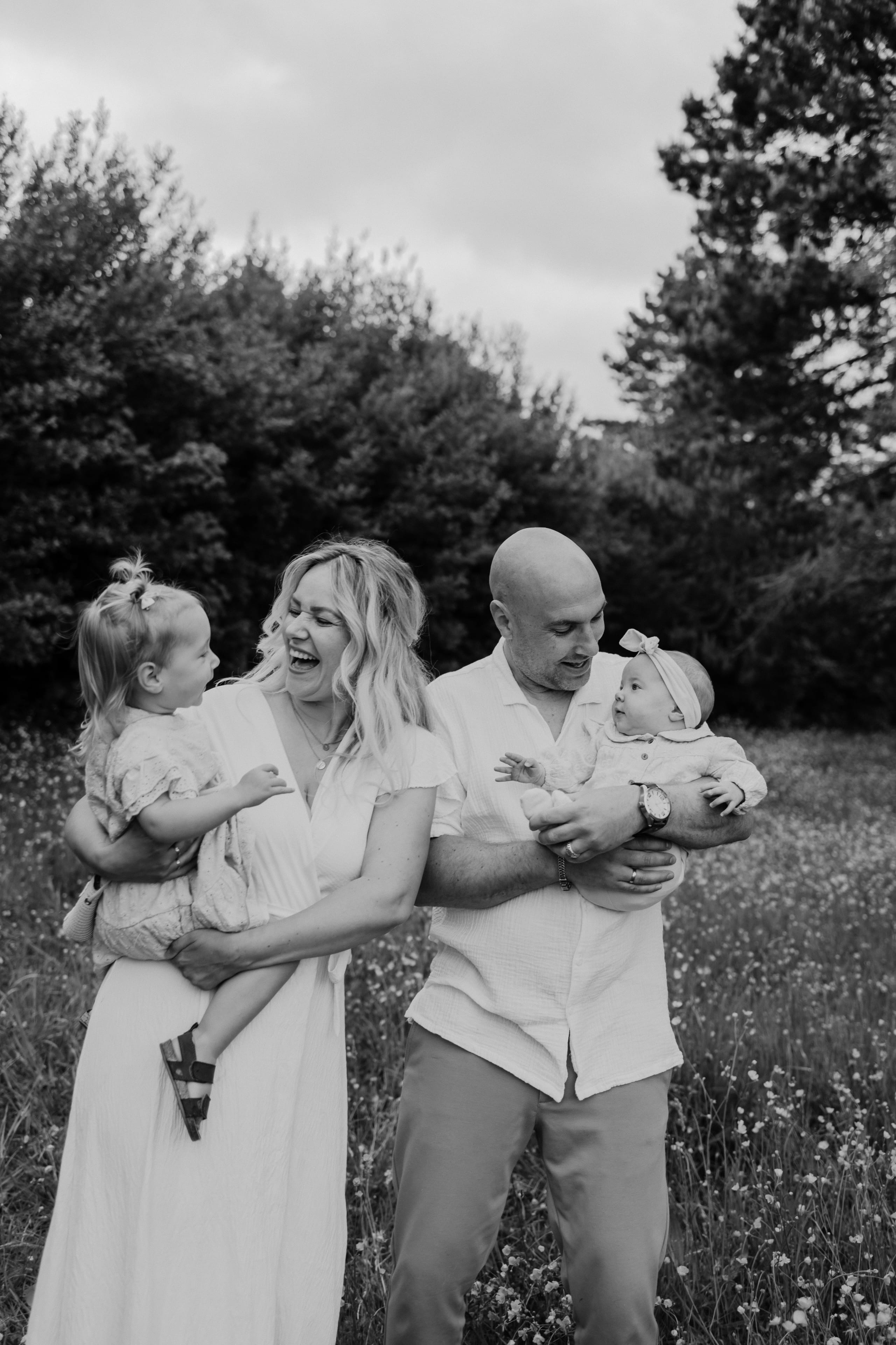 Een gelukkige familie staat in een bloeiende weide, waarbij een vrouw met een witte jurk and a man met een lichtgekleurde blouse samen met twee jonge kinderen op schoot lachen en genieten van het moment.
