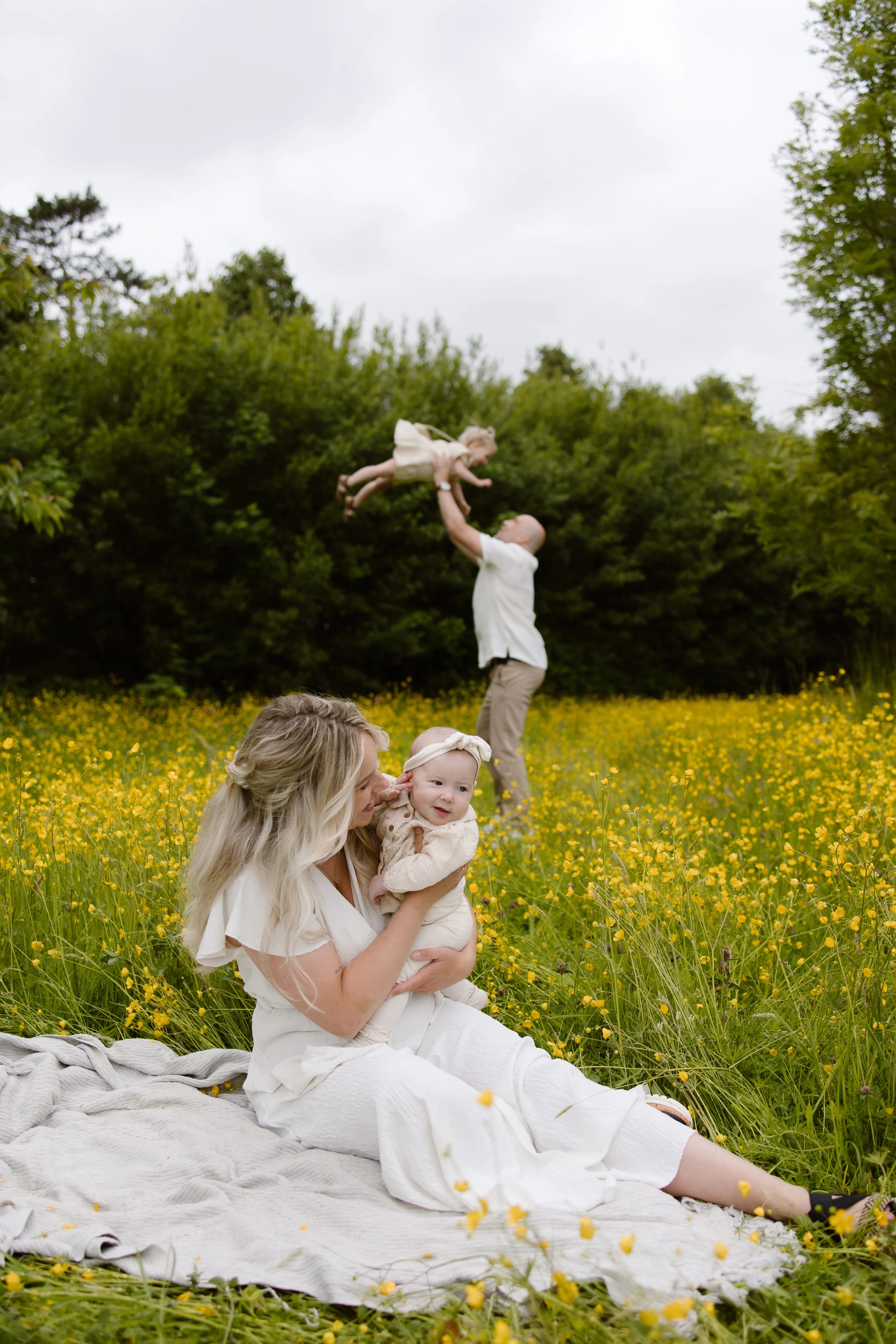 Een moeder knuffelt haar baby op eendeken in een met gele bloemen bedekte weide, terwijl een vader hun oudere kind omhoog tilt, wat een krachtig en vrolijk gezinsmoment vastlegt.