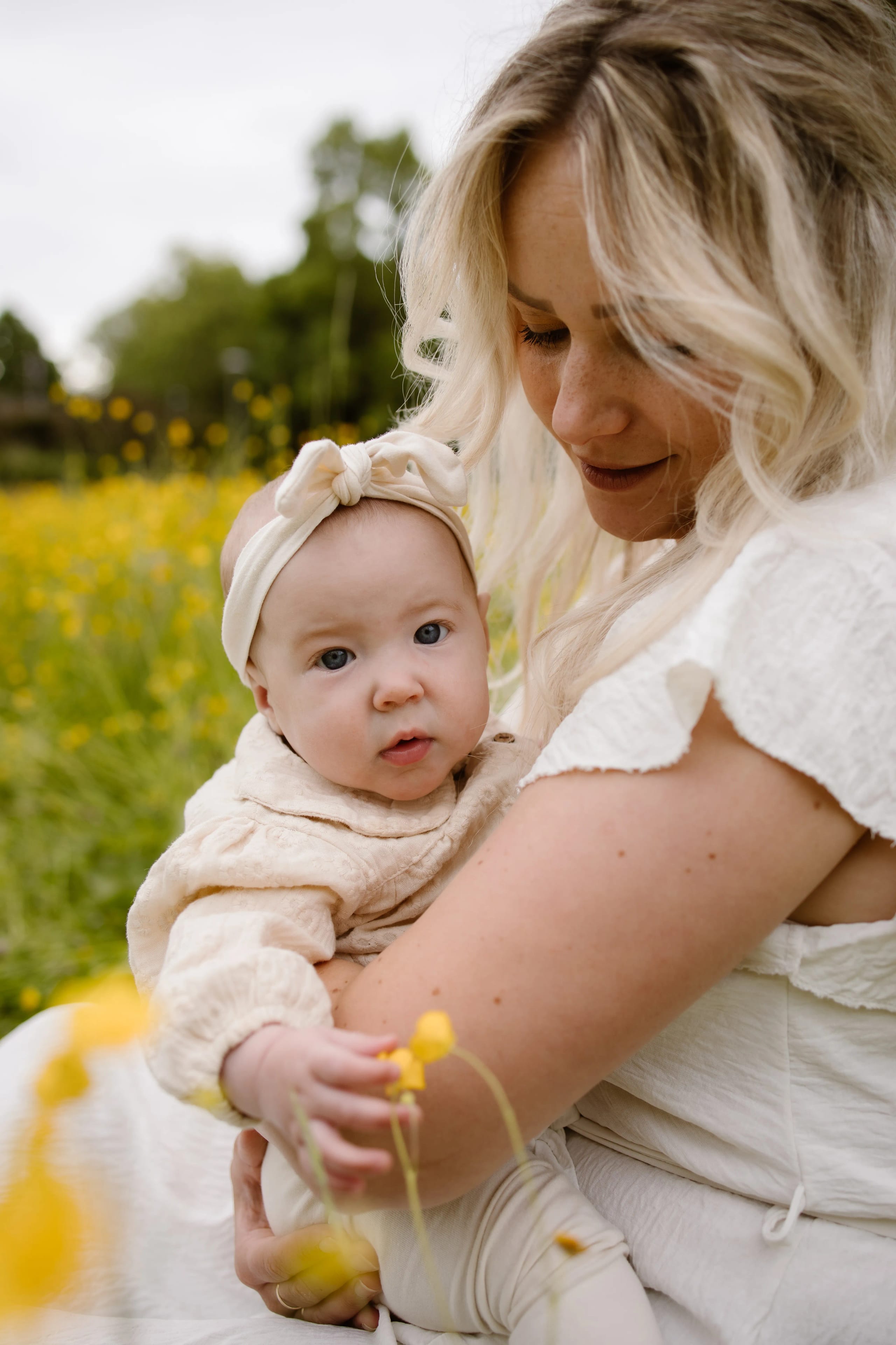 Een moeder houdt haar baby in haar armen terwijl ze in een bloeiend veld met gele bloemen zitten; the baby kijkt nieuwsgierig met een schattige haarband en een lichte trui aan.