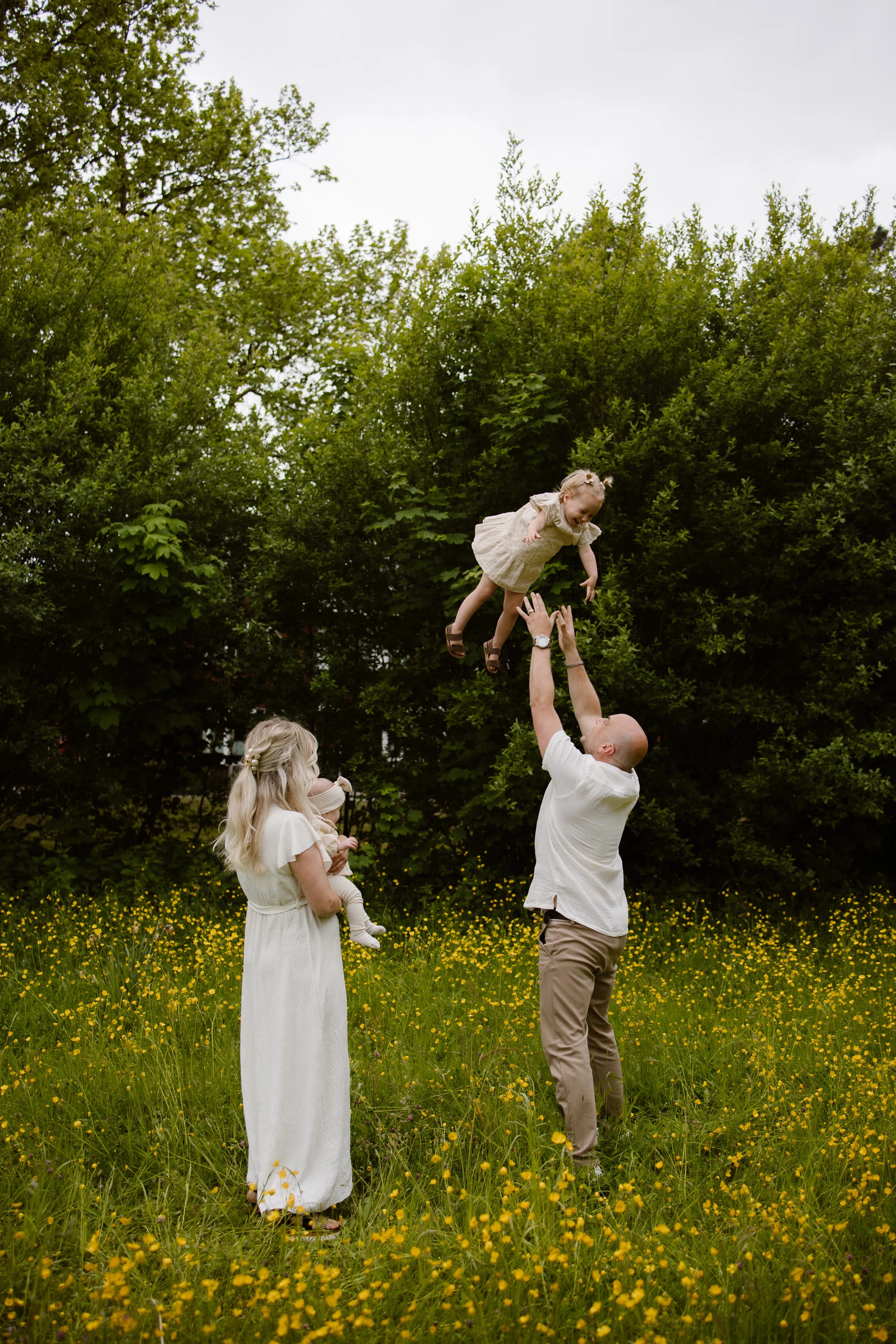 Een vrolijke familie buitenshuis waarbij een vader zijn jonge dochter in de lucht gooit terwijl de moeder een baby vasthoudt, omringd door een groene omgeving en gele bloemen.