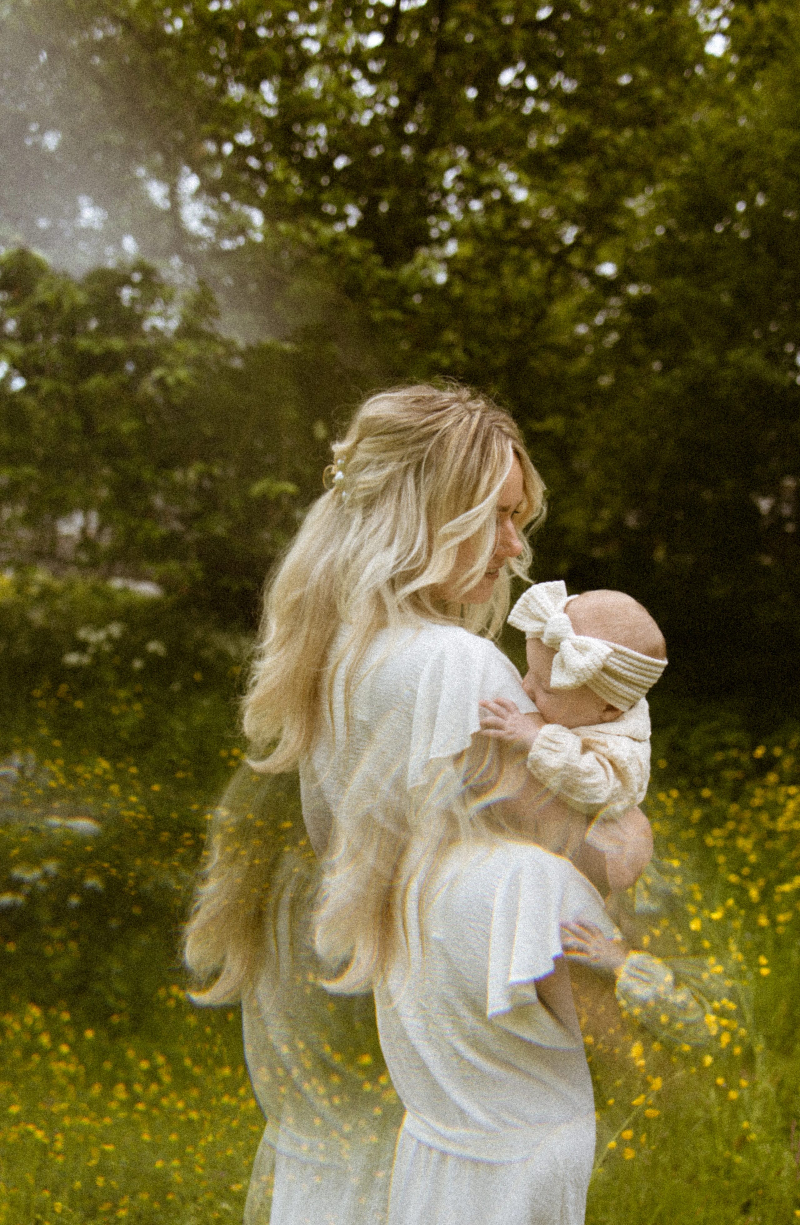 Een moeder houdt haar baby met een schattige haarband vast, terwijl ze in een bloemenveld staat, omringd door groene bomen en een dromerige sfeer.