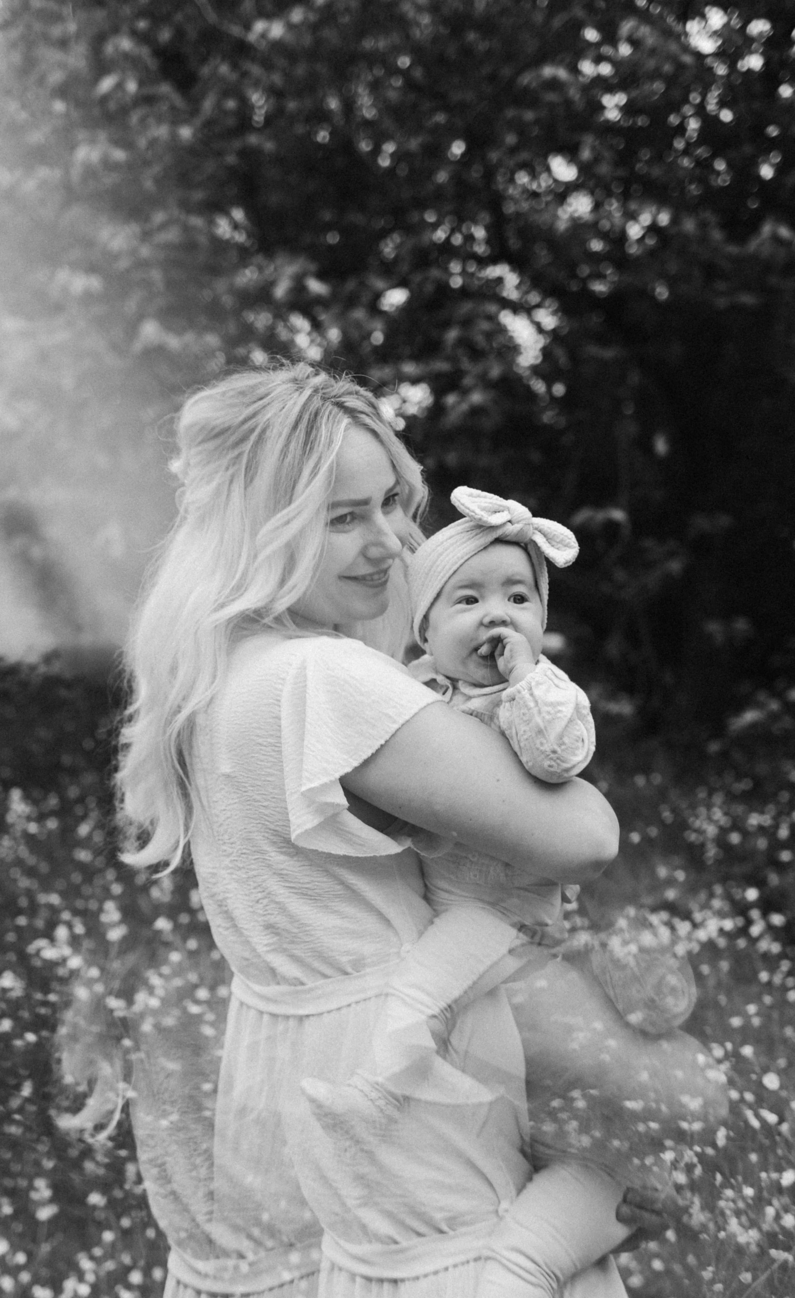 Een moeder met lang, golvende haar houdt liefdevol een baby vast die een schattig haarbandje draagt, terwijl ze samen in een bloeiende, natuurlijke omgeving staan. De afbeelding straalt een gevoel van warmte en verbondenheid uit.