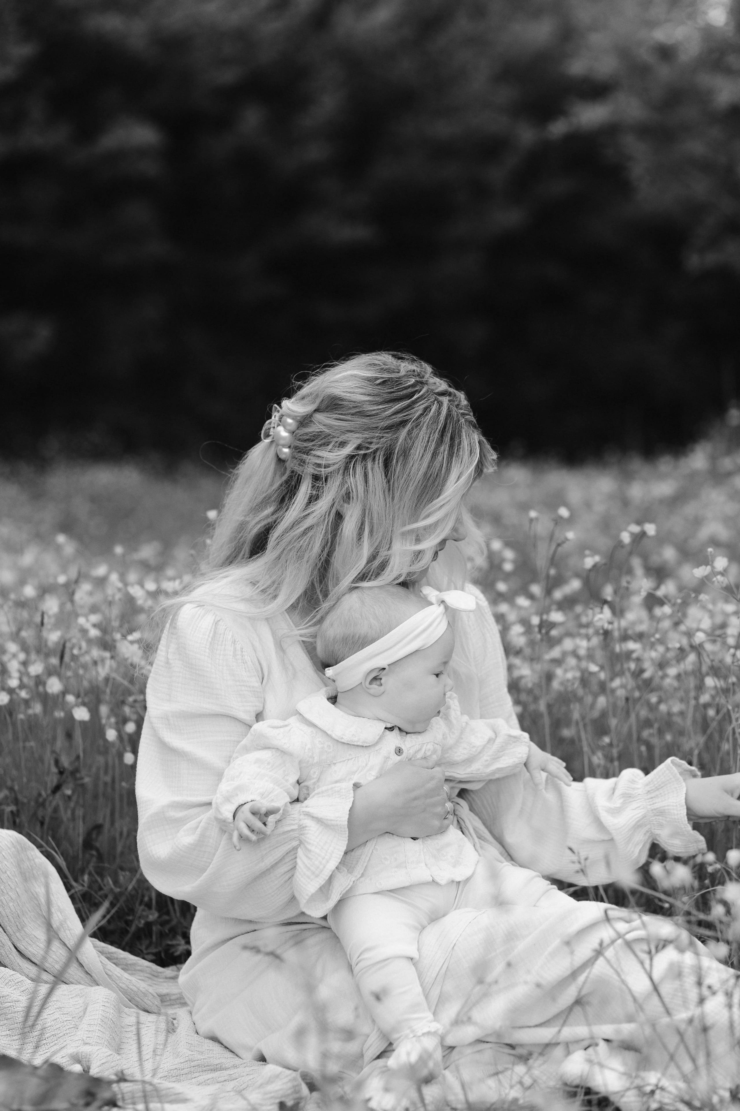 Moeder en baby zitten samen op een kleed in een bloeiend grasveld, waarbij de moeder liefdevol naar de baby kijkt die een schattige hoofdband draagt.