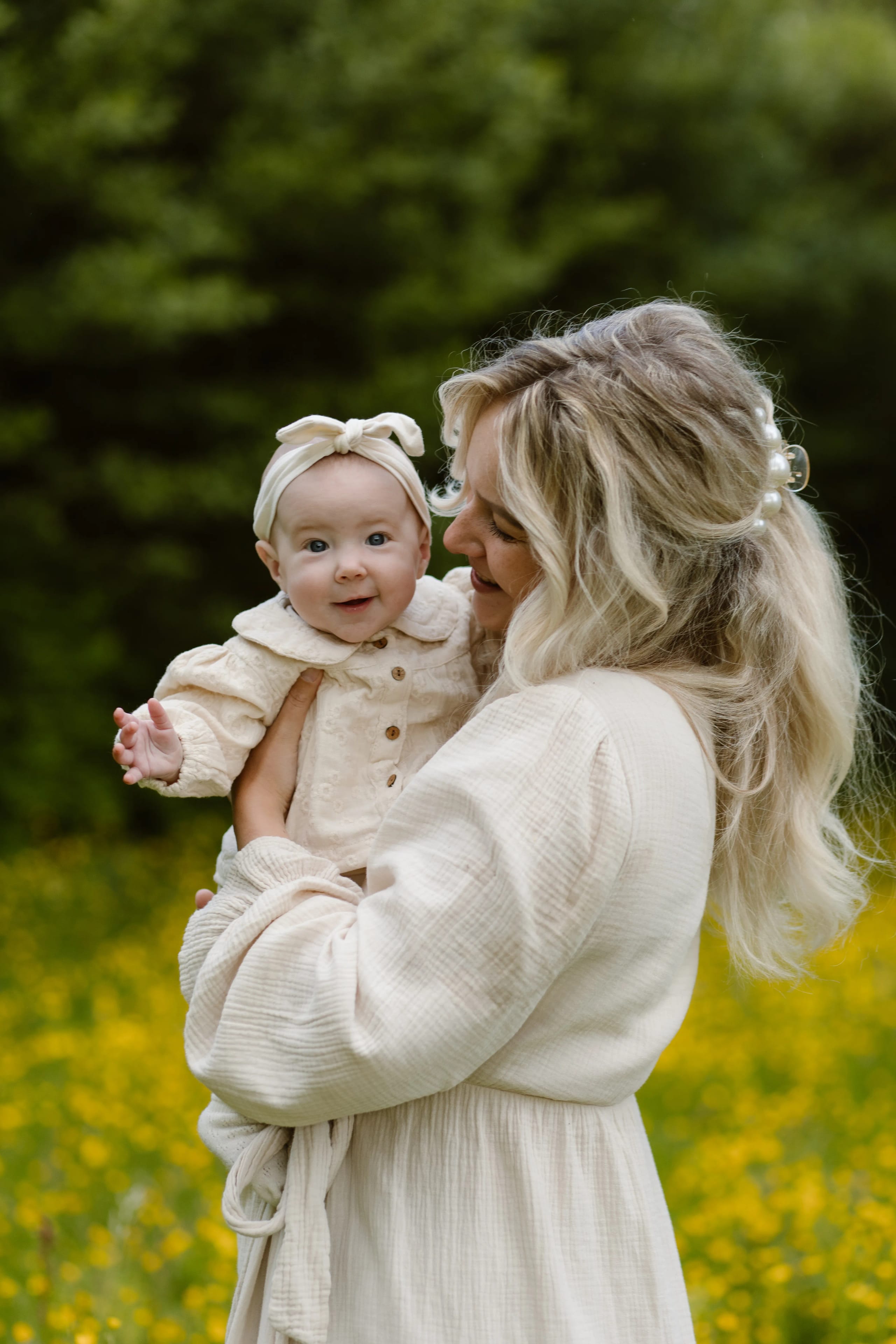 Een blije moeder houdt haar schattige baby in een groene, bloemenrijke omgeving vast, terwijl ze elkaar liefdevol aankijken. De baby draagt een schattige haarband en de moeder is gekleed in een stijlvolle, lichte outfit.