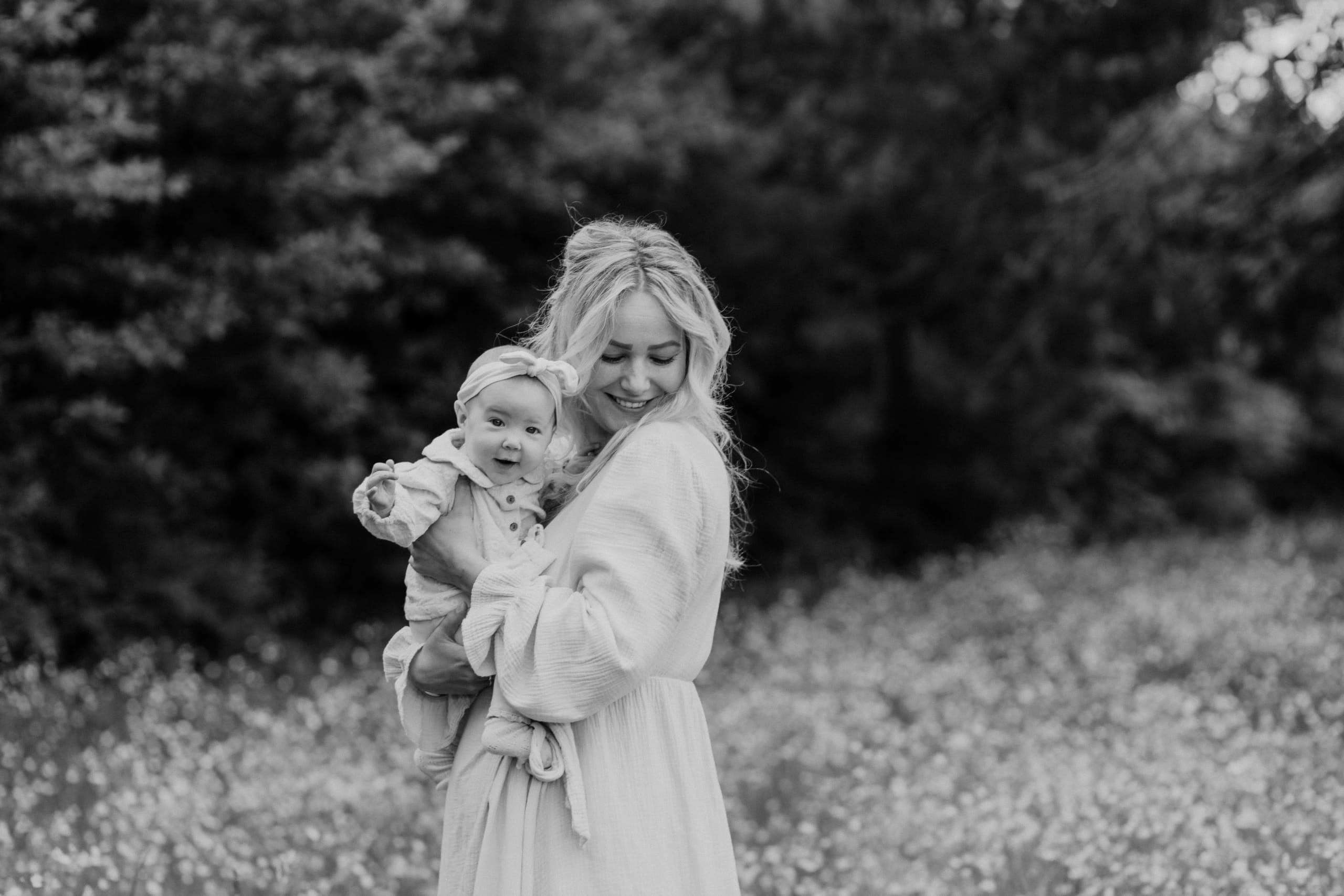 Vrouw in een lichte, vloeiende jurk houdt blije baby in een bloemrijke, groene omgeving vast, met een warme glimlach op haar gezicht.