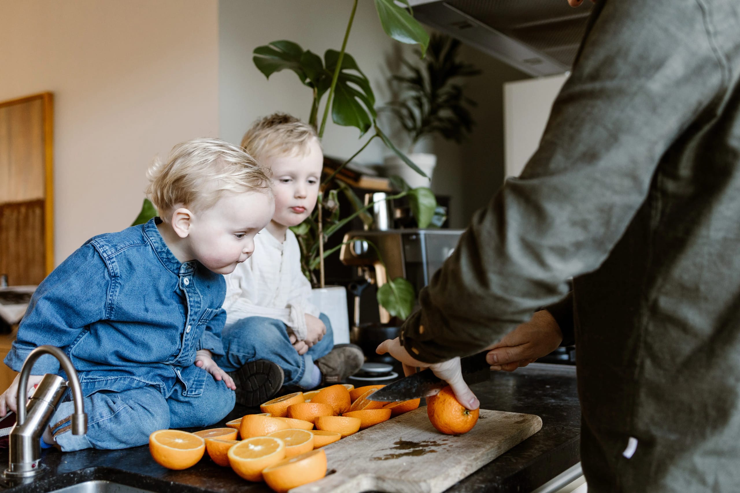 Twee jonge kinderen kijken toe terwijl hun vader fruit snijdt in de keuken, authentieke lifestyle fotografie.