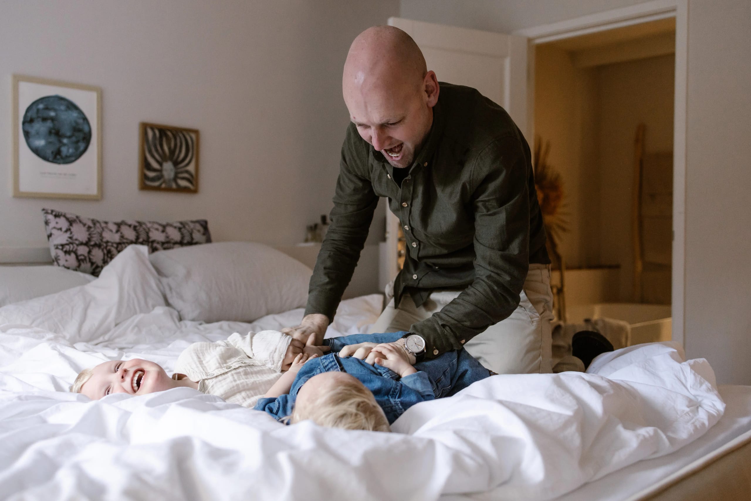 Vader kietelt zijn zoontje op bed, echte emoties tijdens een familie fotoshoot thuis.