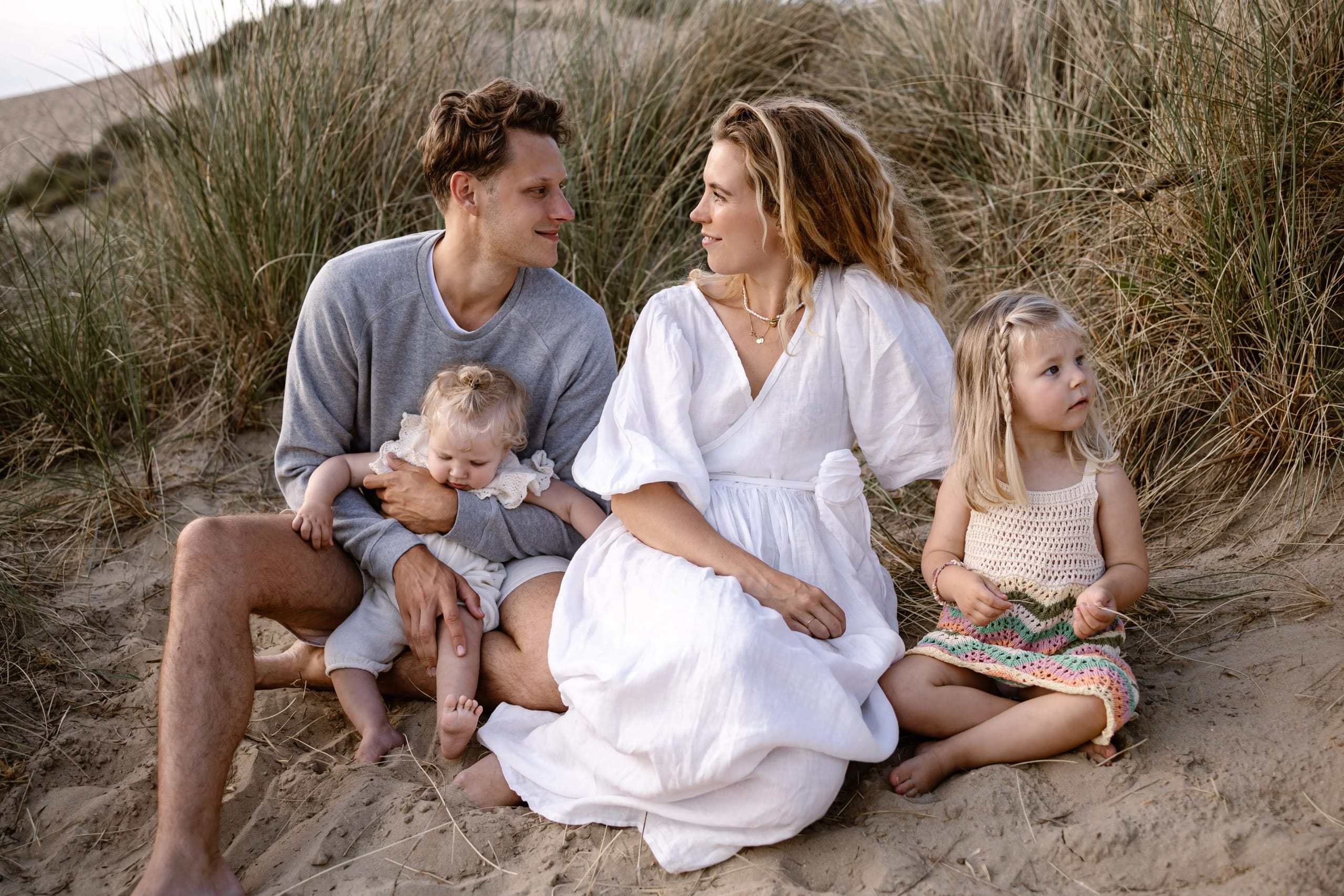 Vader, moeder en kinderen zitten samen in het zand van de Bloemendaalse duinen, ontspannen familieportret.