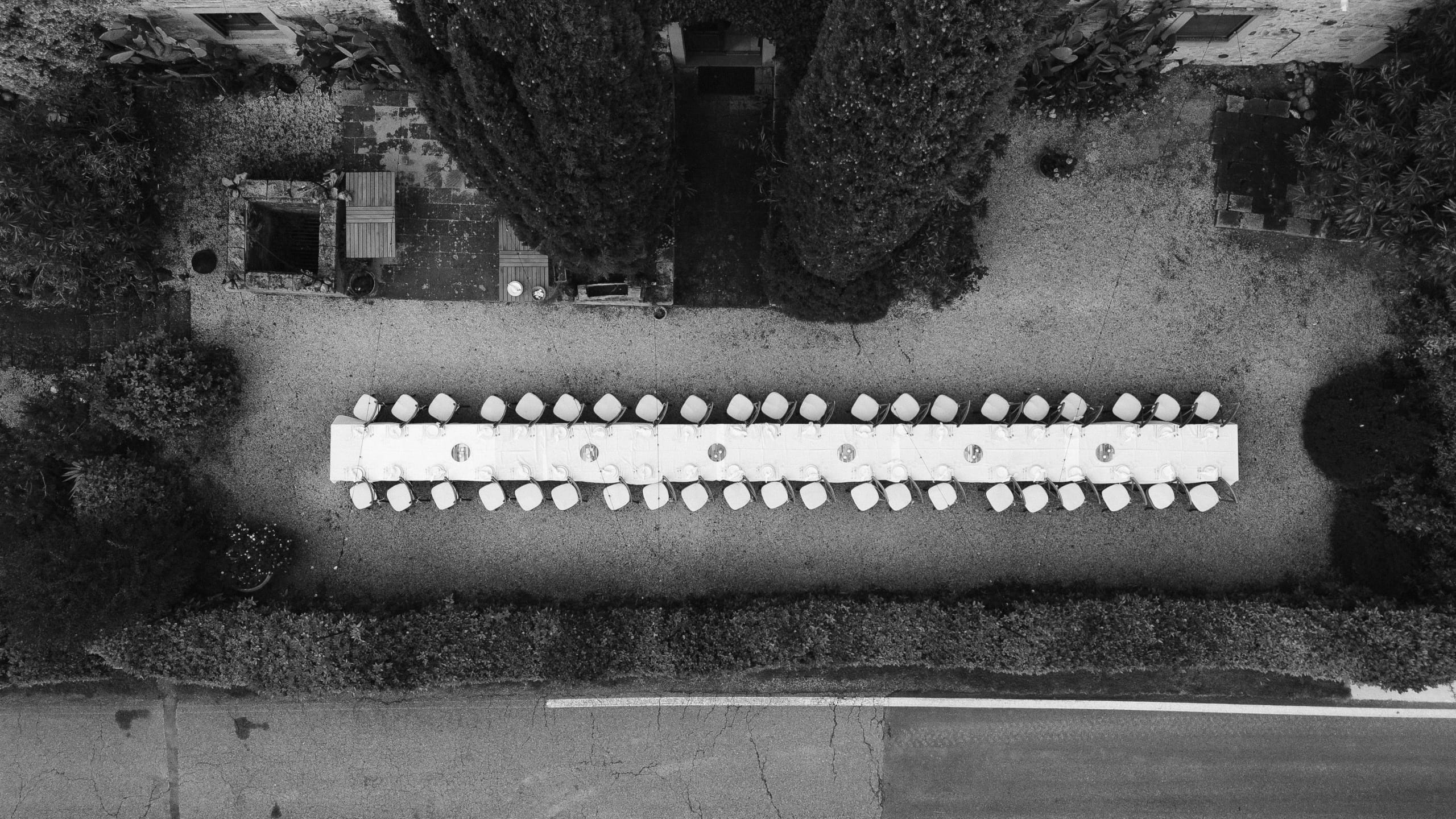 Langwerpige eettafel met witte tafelkleden en stoelen, omringd door weelderige groene planten en een grindpad, gefotografeerd vanuit de lucht in een serene buitenomgeving.