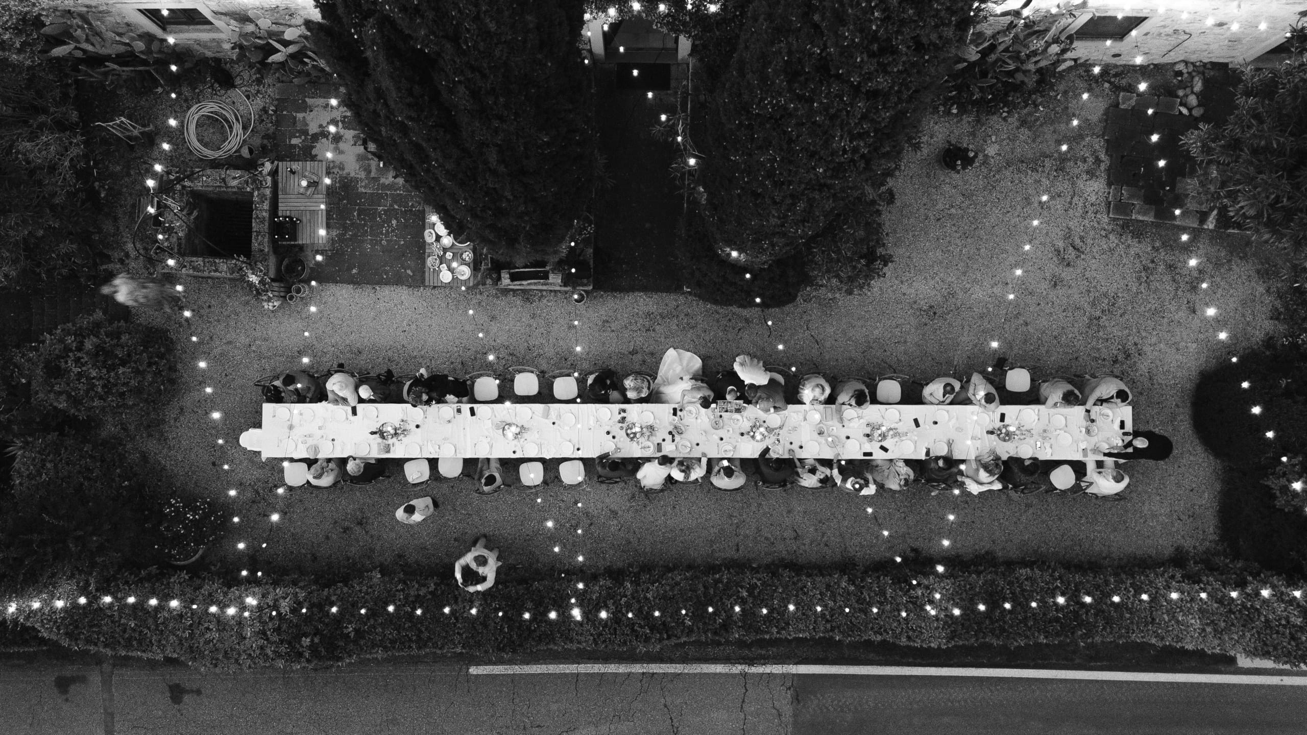 Een overzicht van een buitenfeest met een lange tafel vol gasten, sfeervol verlicht door twinkelende lichtjes en omringd door weelderige groene planten, waarbij mensen genieten van het samenzijn en de gezelligheid in een tuinsetting.