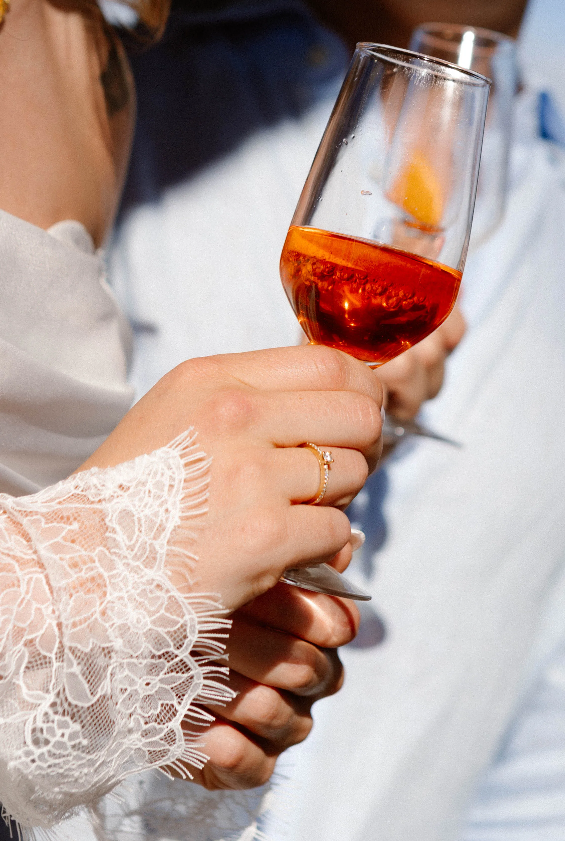 Een hand met een delicate, kanten mouw holding een glas met een oranje drankje, terwijl op de achtergrond een andere persoon aanwezig is.