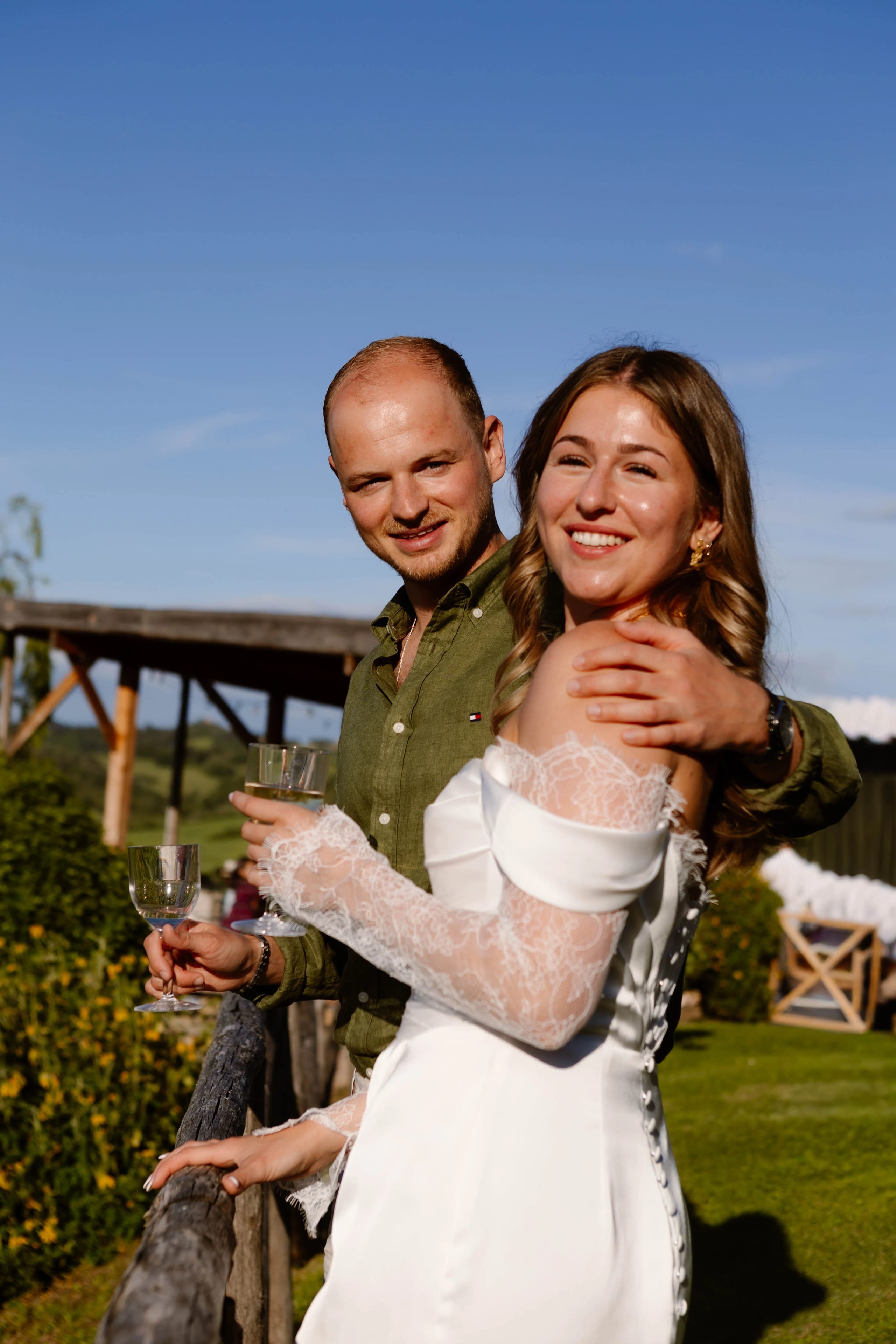 Een gelukkige jongeman en vrouw, gekleed voor een bruiloft, poseren liefdevol samen met glazen in hun handen, omgeven door een schilderachtige groene omgeving en een heldere blauwe lucht.