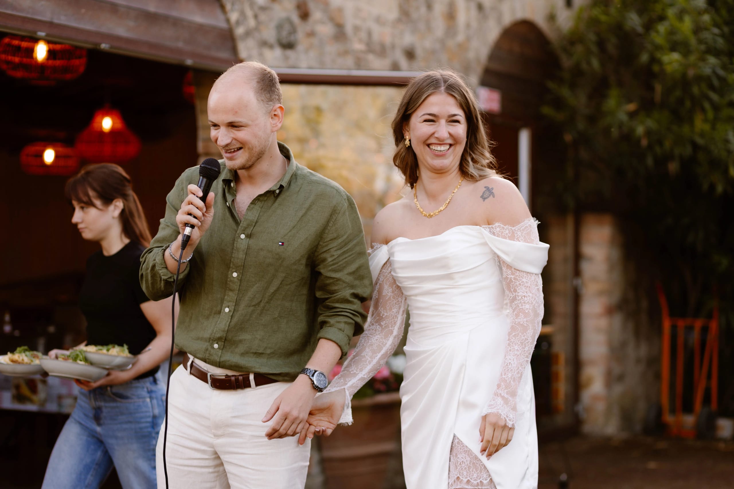 Een man en een vrouw staan hand in hand op een stijlvolle buitenlocatie, terwijl de man een microfoon vasthoudt en naar het publiek spreekt. De vrouw, gekleed in een elegante witte jurk, glimlacht breed, en op de achtergrond is een serveerster te zien met een dienblad vol salades.