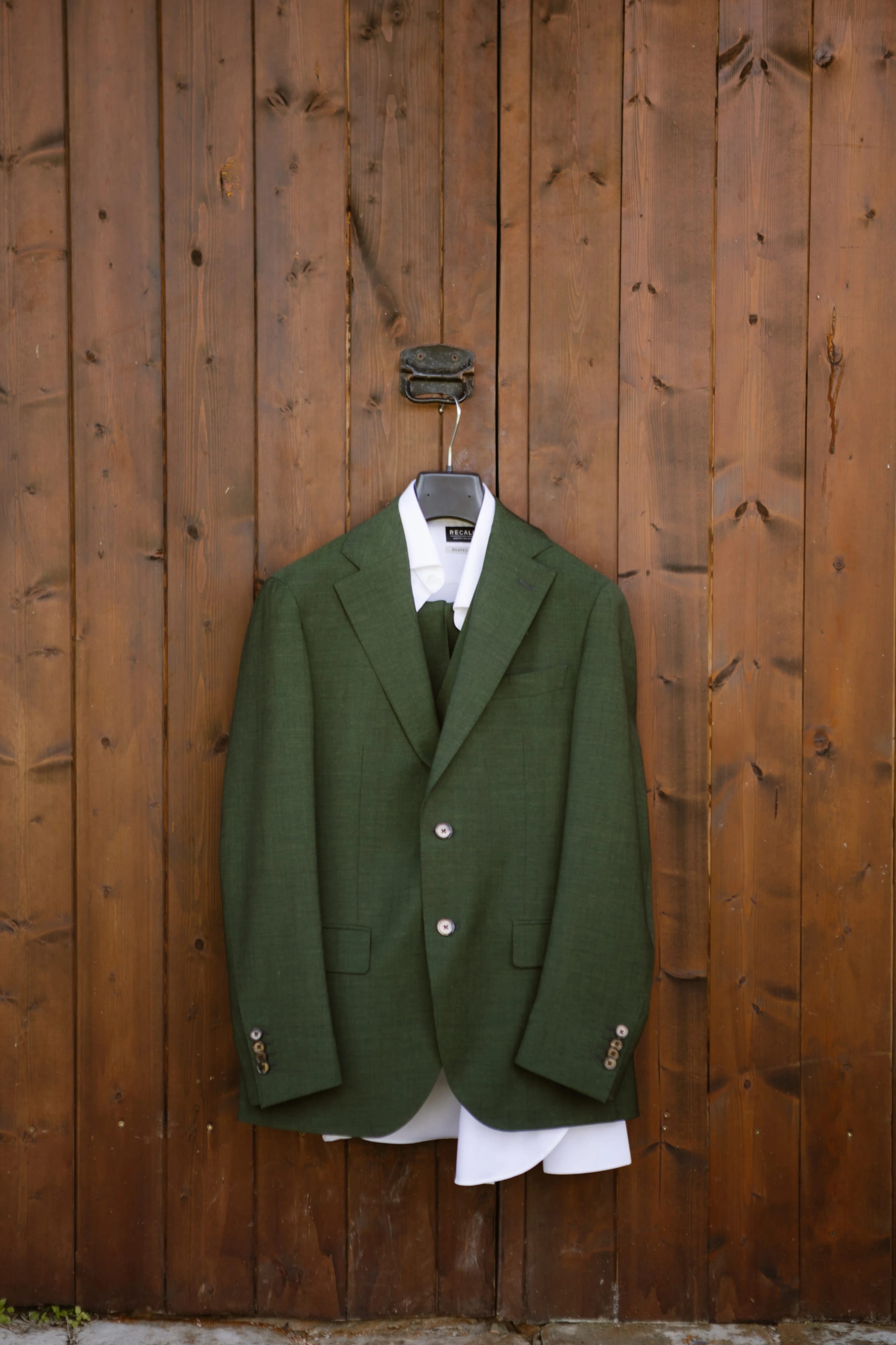 Groene blazer met bijpassend vest en witte overhemd, elegant opgehangen aan een hanger voor een houten achtergrond.