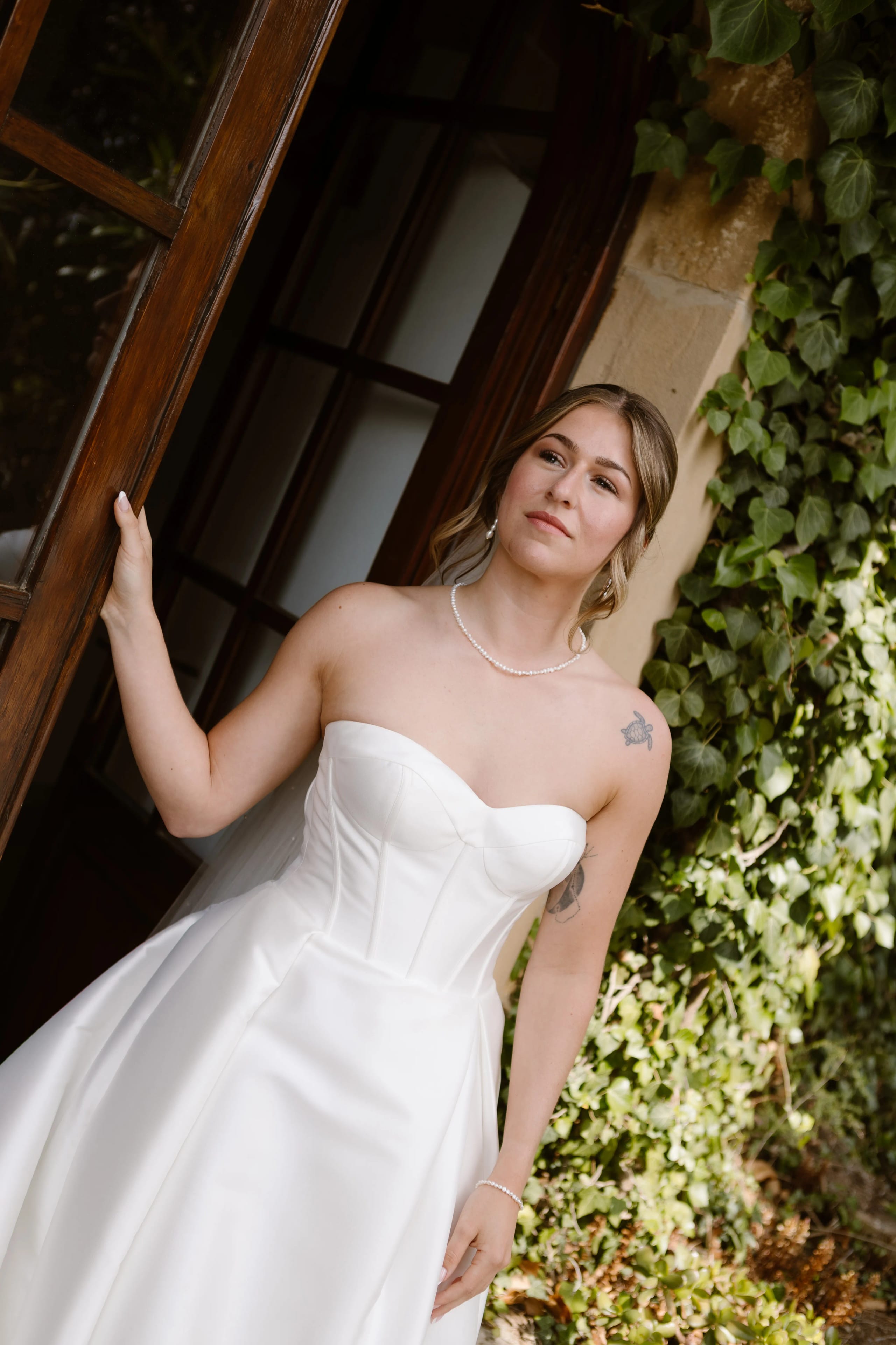 Een bruid in een strapless, zijden trouwjurk poseert elegant bij een houten deur, omringd door groene klimop, met opgestoken haar en smaakvolle parelaccessoires die haar uitstraling verfijnen.