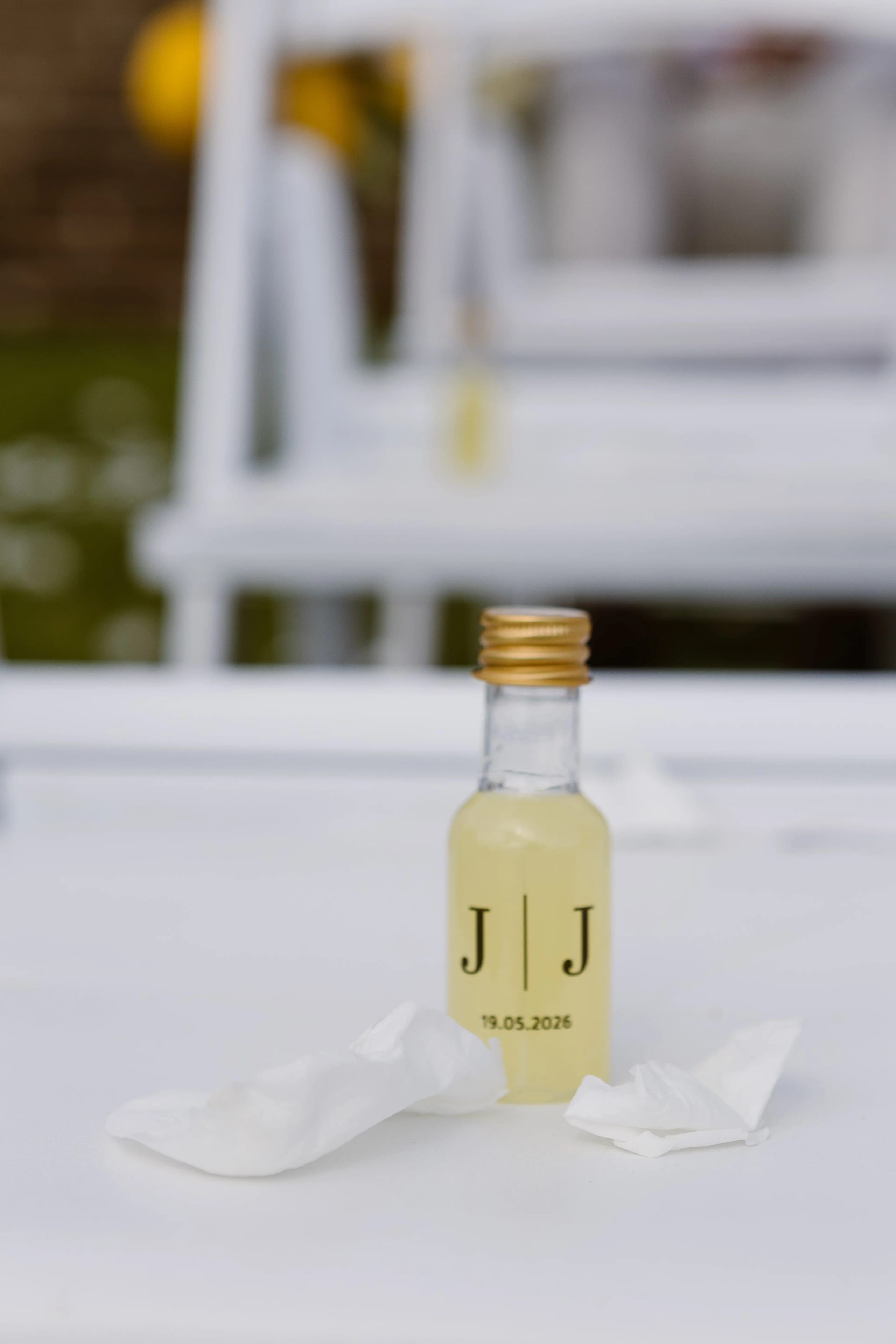 Klein glasflesje met een lichte drank en een gouden dop, voorzien van een zwart logo en een datum, ligt op een witte tafel, omringd door een paar crumpled servetten, wat suggereert dat het evenement speciaal of feestelijk is.