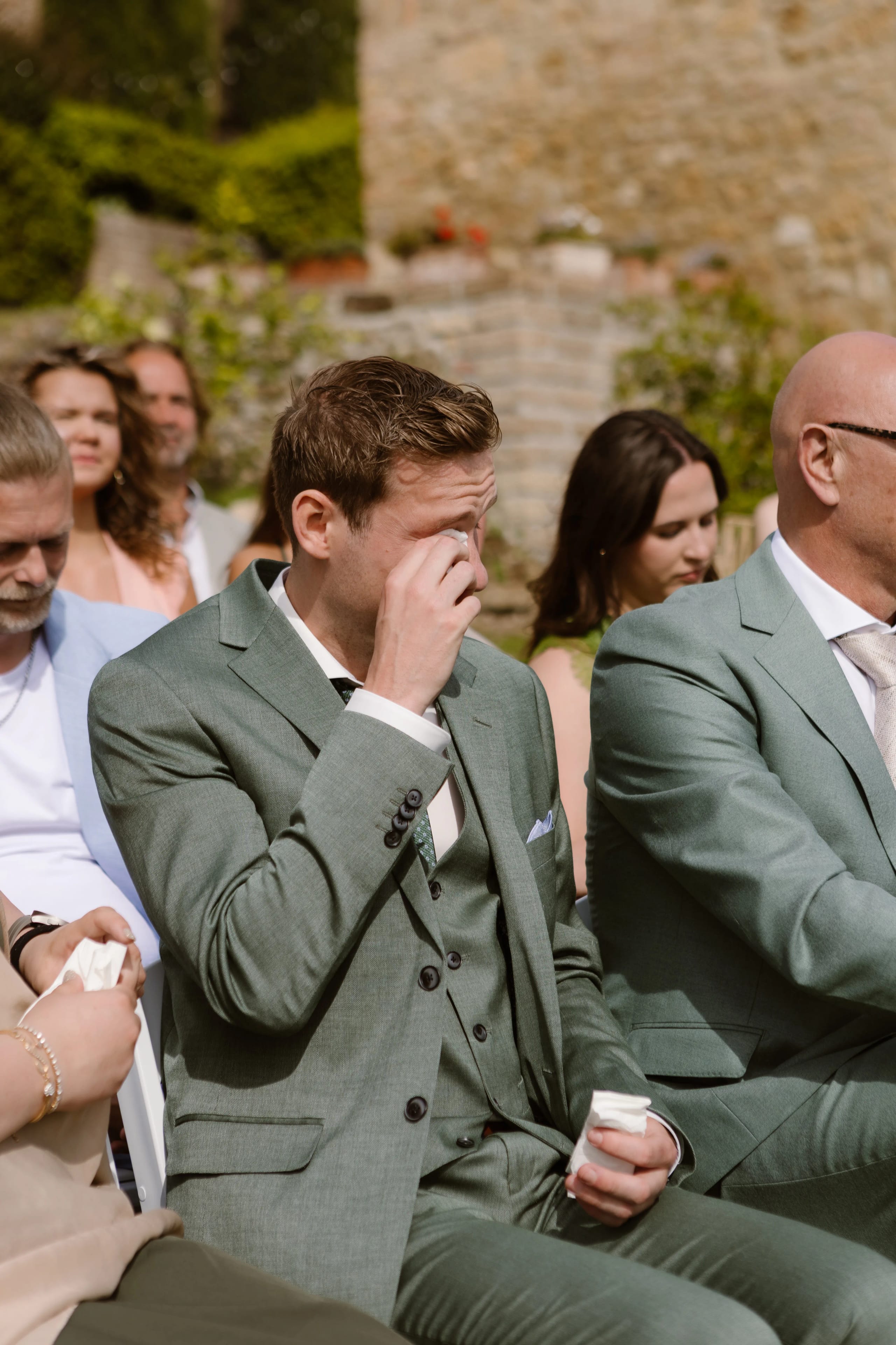 Emotionele bruiloftsmoment met een jongeman in een groene pak die zijn tranen wegveegt, omringd door een getuigenis van vrienden en familie in een prachtige buitenomgeving.