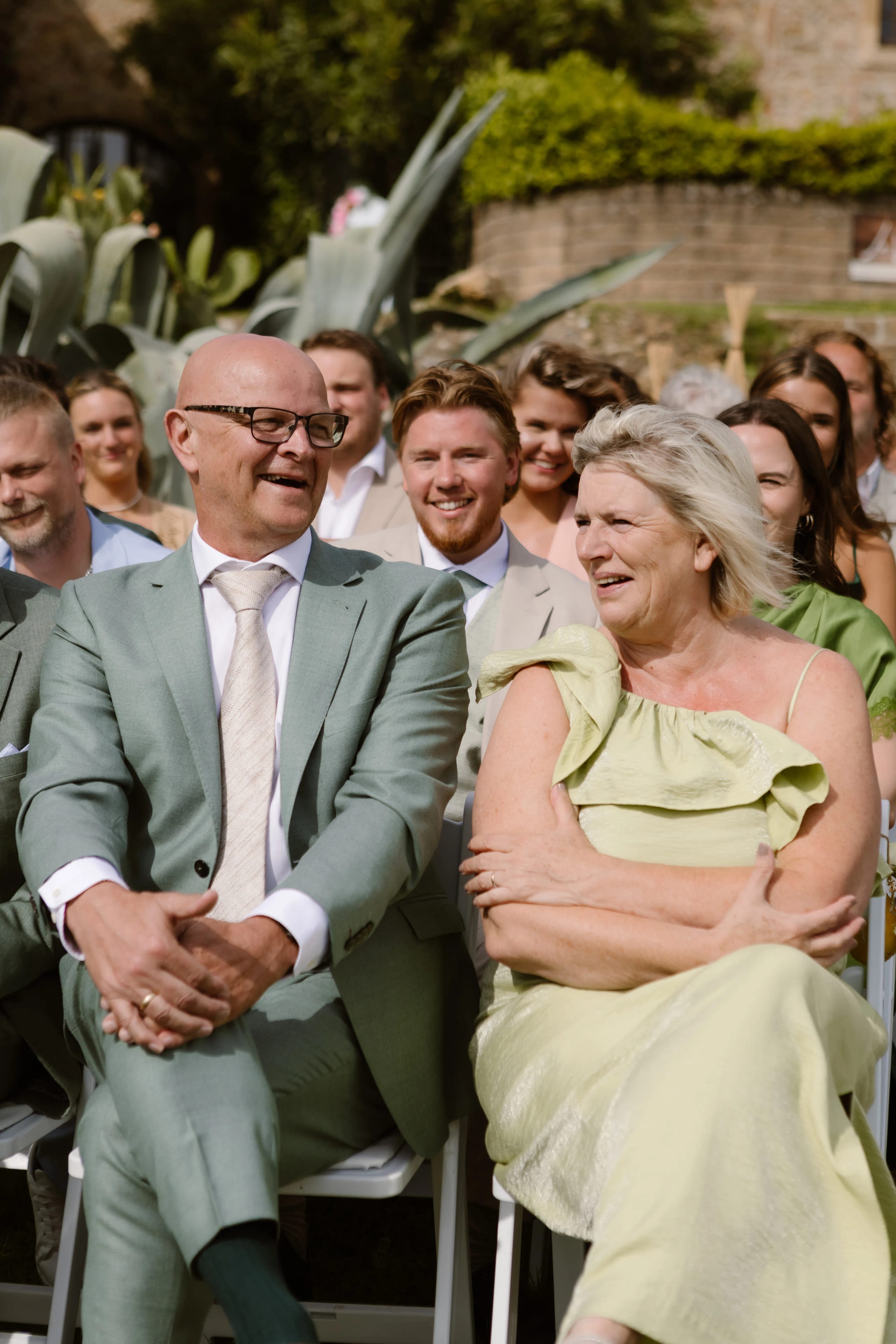 Een stijlvol gekleed echtpaar met een ontspannen houding, zittend op een ceremonie, omringd door vrienden en familie, met groene planten op de achtergrond. De vrouw in een groene top lacht terwijl de man in een lichtgroen pak haar aandacht vasthoudt.
