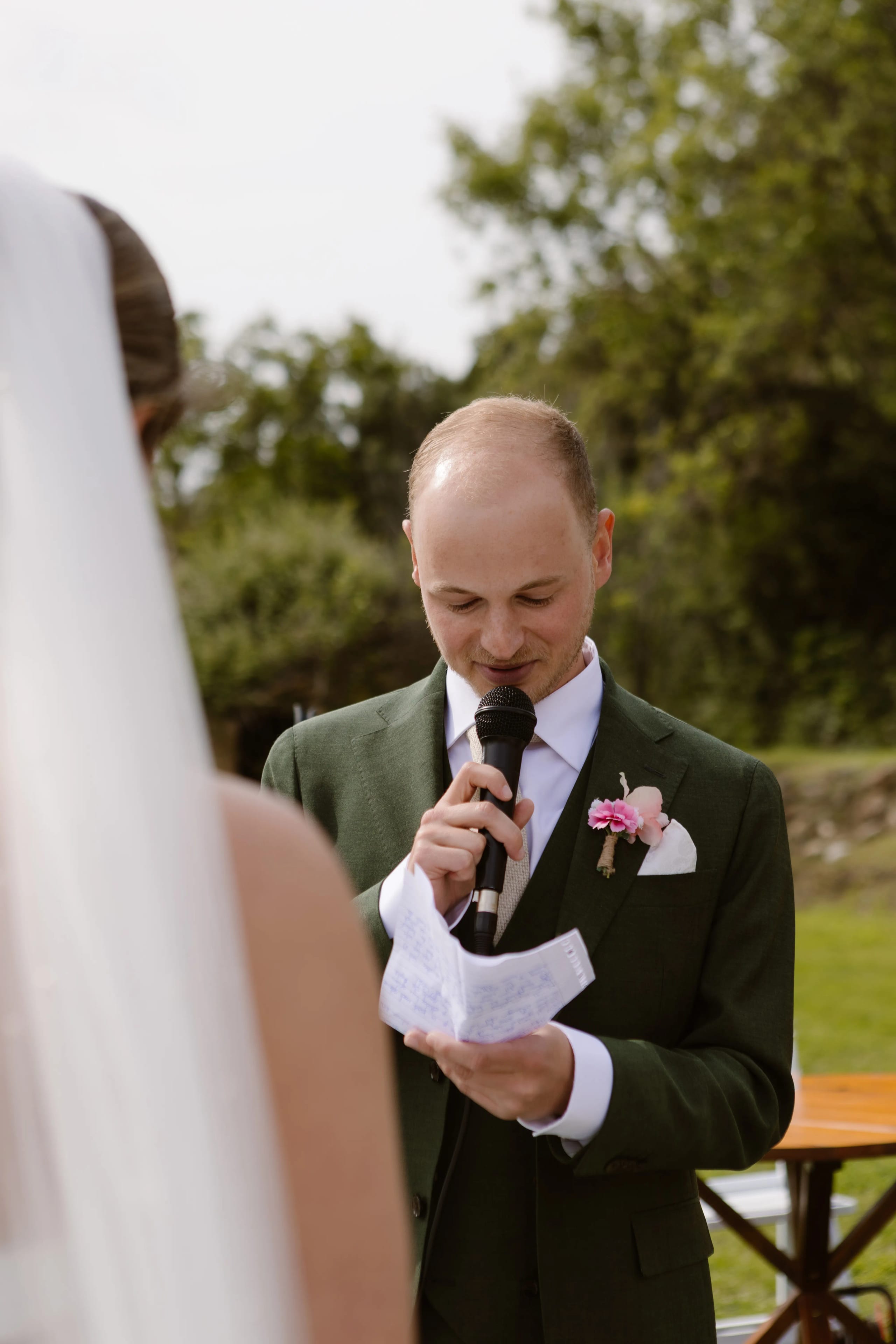 Een man in een groen pak houdt een microfoon vast terwijl hij een tekst voorleest tijdens een huwelijk, met op de achtergrond een vrouw in een witte trouwjurk en een groene, natuurlijke omgeving.