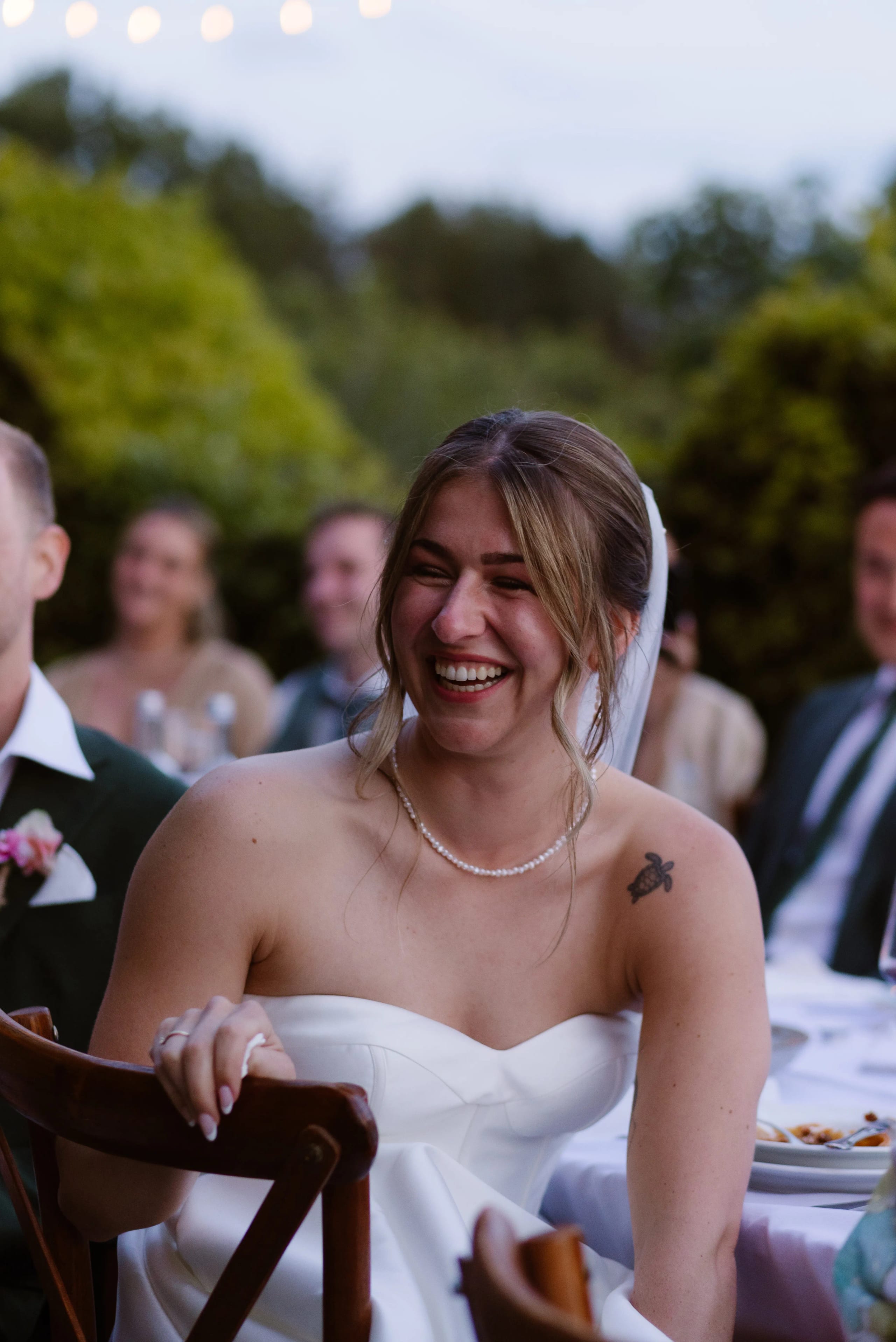 Een bruid met een stralende lach in een strapless trouwjurk, genietend van een vrolijk moment tijdens de huwelijksreceptie, omringd door gasten en een groene tuin op de achtergrond.