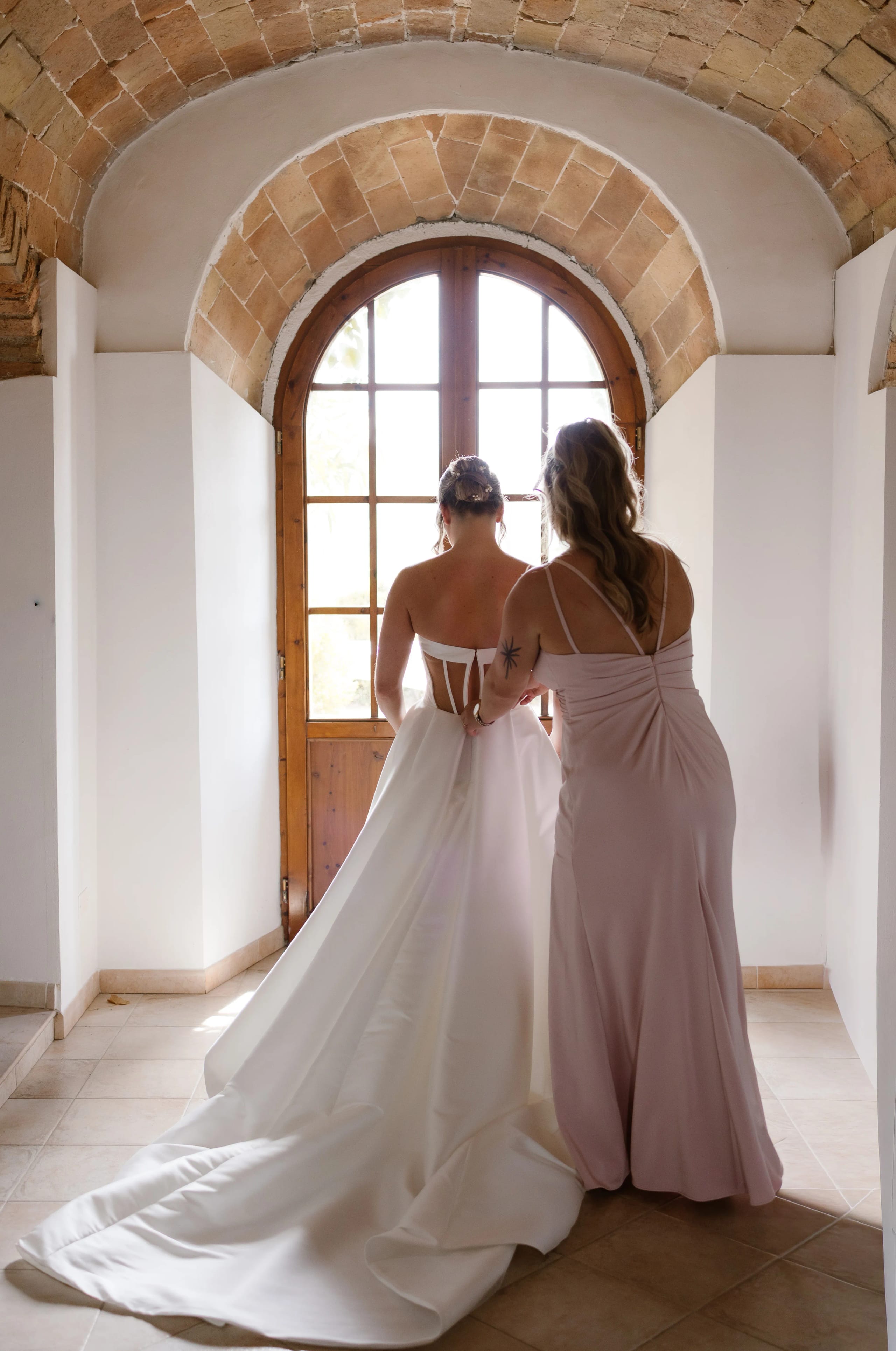 Een bruid in een strapless witte trouwjurk wordt geholpen door een vriendin in een lichtroze jurk, terwijl ze naar een grote houten deur met een gebogen boog kijkt, wat een feestelijke sfeer voor hun speciale dag creëert.