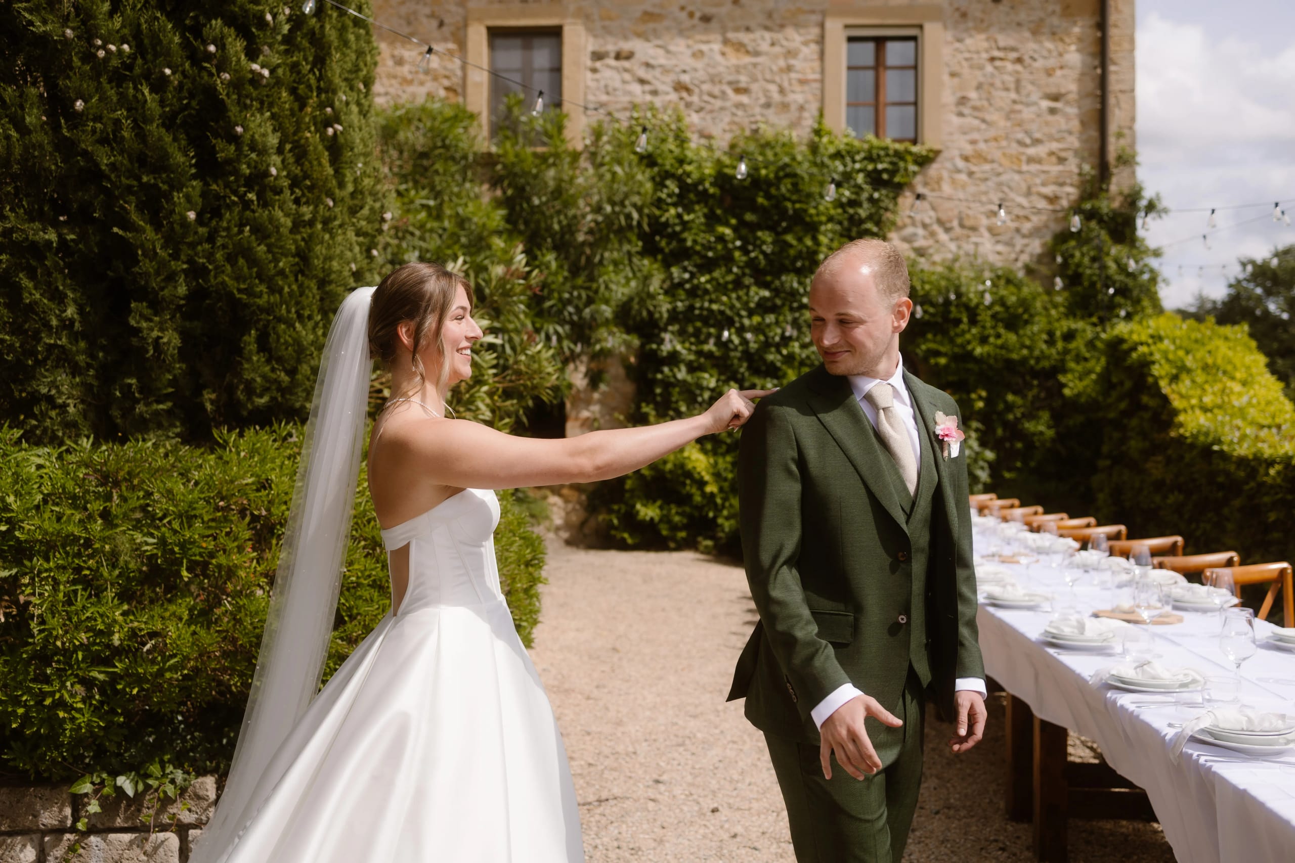 Een bruid in een elegante witte jurk en een bruidegom in een groen pak lachen naar elkaar terwijl ze op een romantische locatie buiten staan, omringd door groen en een mooi gedekte tafel voor een huwelijksreceptie.