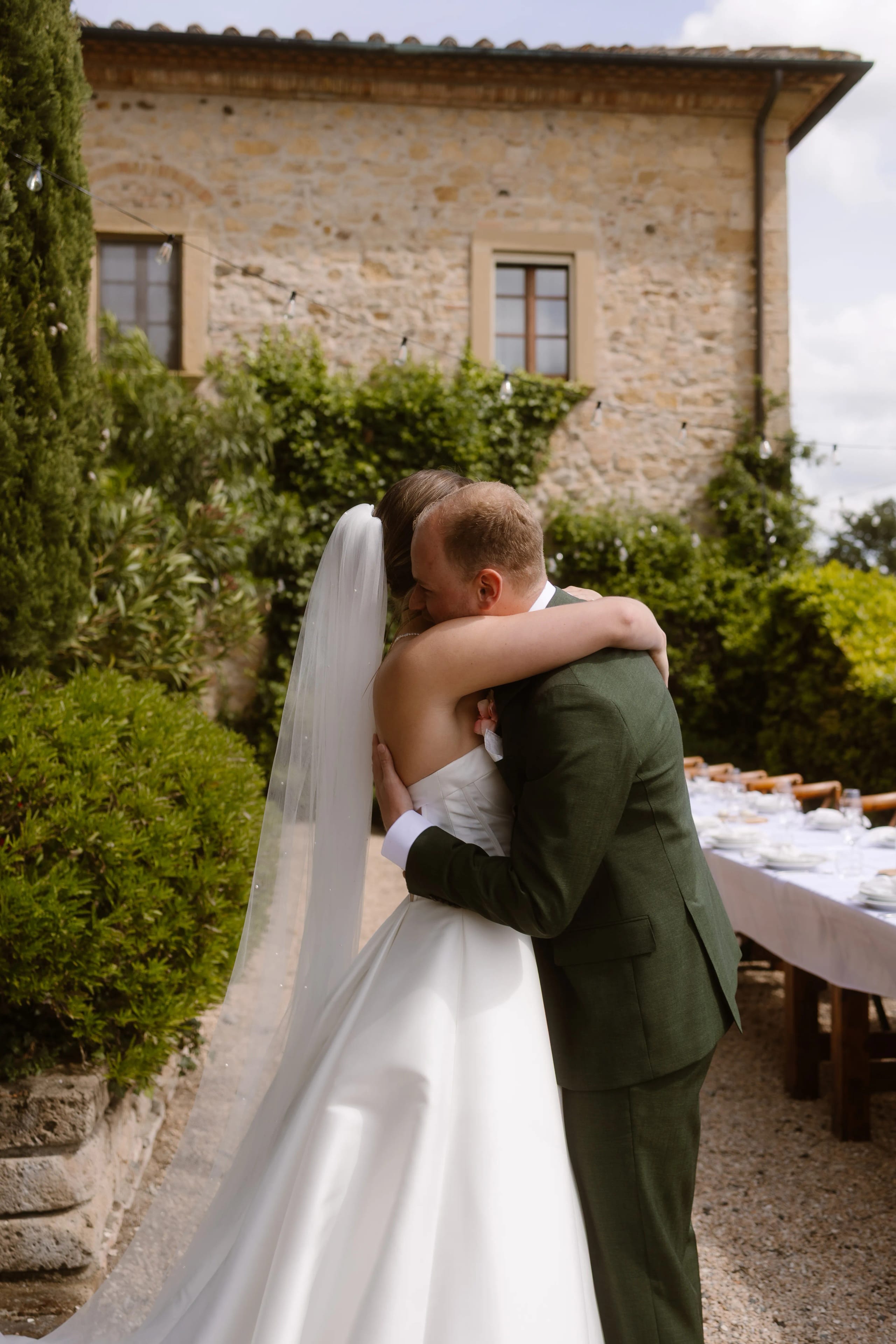 Een pasgetrouwd stel omarmt elkaar liefdevol onder de warme zon, omringd door een groene tuin en een oude stenen villa, terwijl een feesttafel in de achtergrond klaarstaat voor de viering.