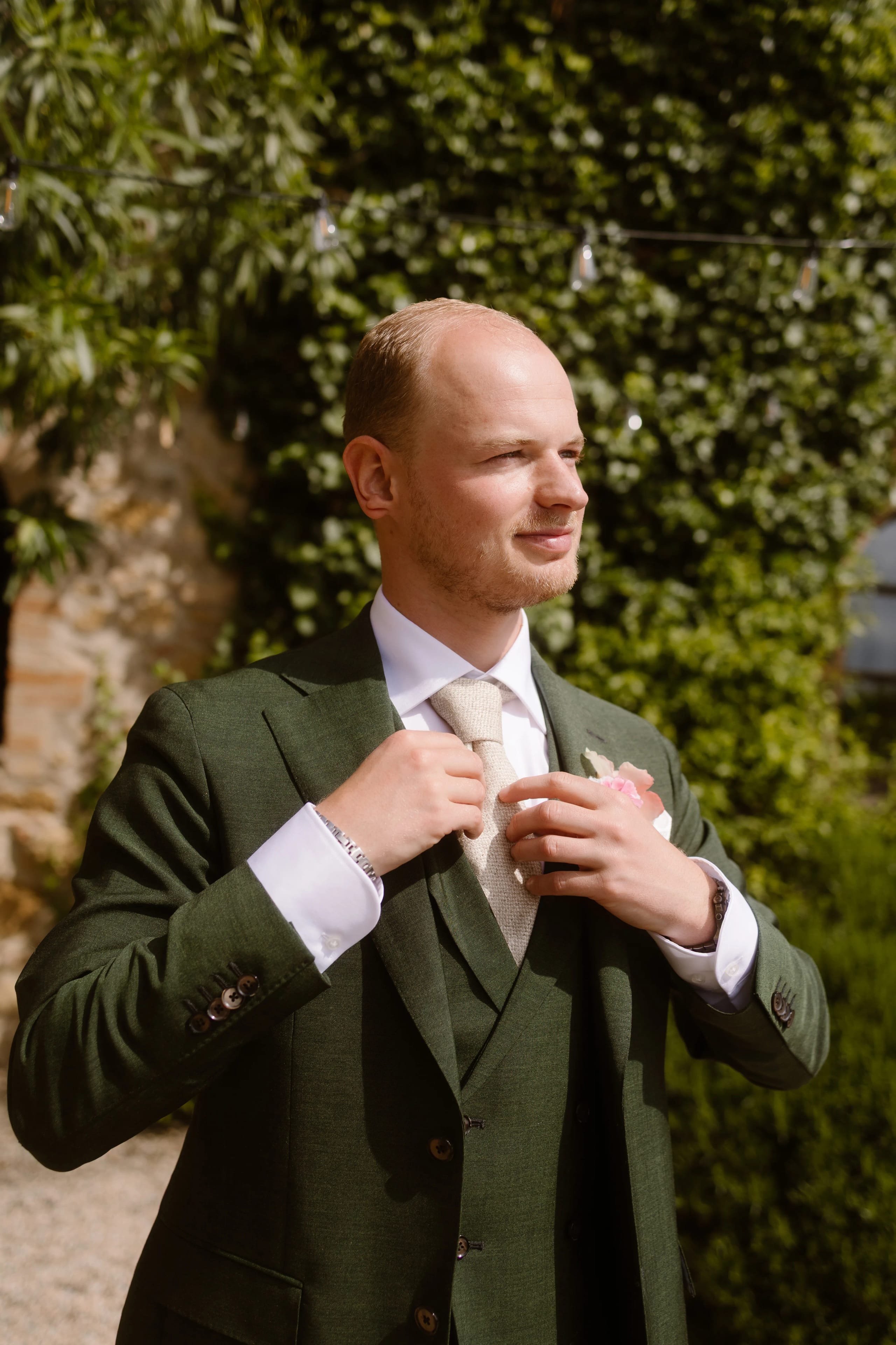 Een man in een groen pak die zijn stropdas strakt terwijl hij geniet van de buitenlucht, omringd door een weelderige groene achtergrond en feestverlichting.