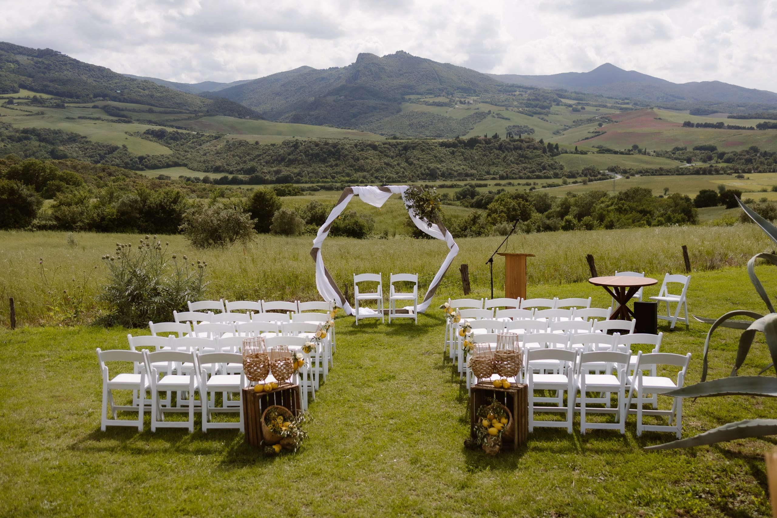 Een romantische buitenceremonie met rijen witte stoelen, een prachtig boogdecor van witte draperieën en bloemen, gelegen in een schilderachtige groene vallei met heuvels op de achtergrond.