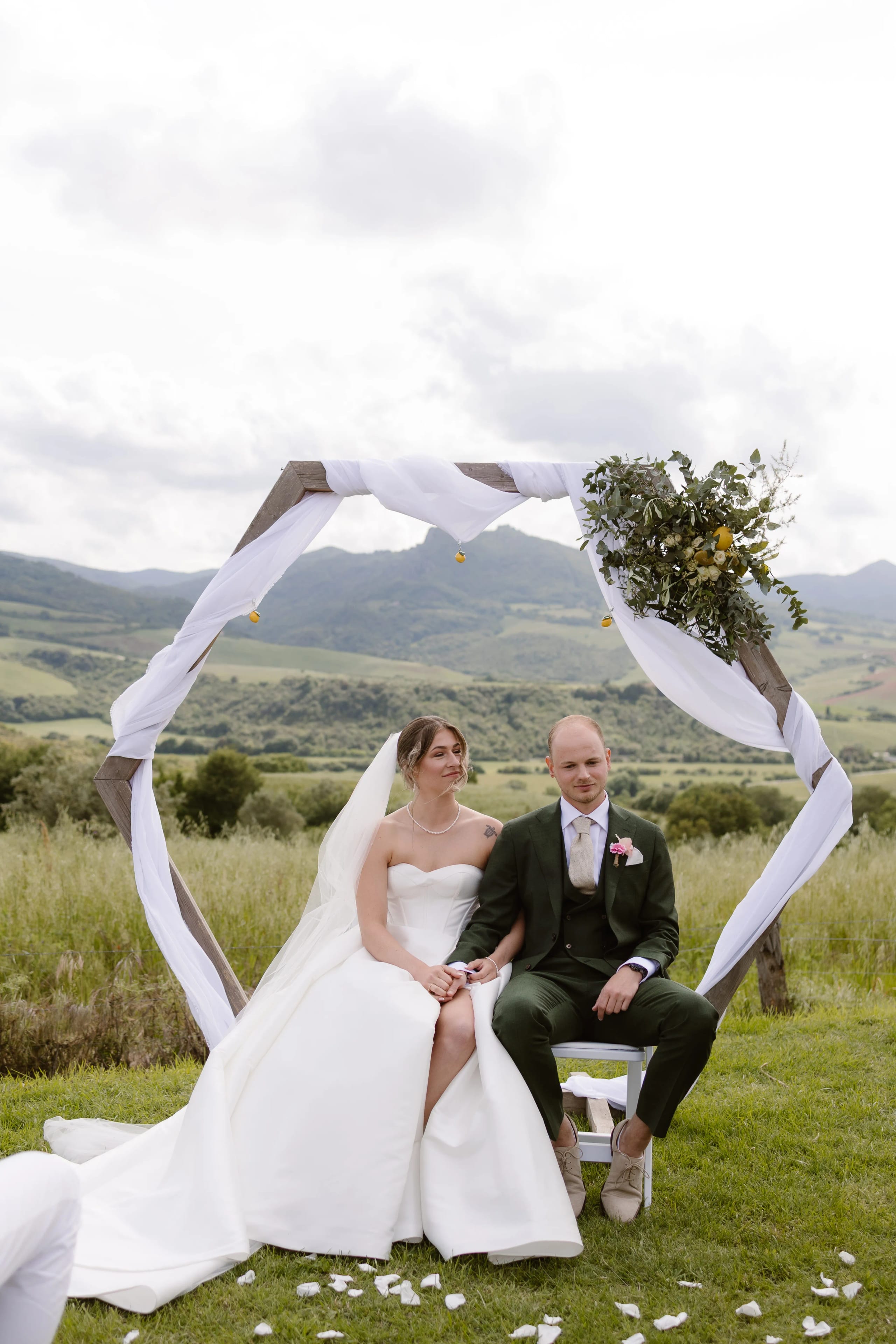 Een bruidspaar zit samen onder een prachtig versierde trouwboog met groene en witte elementen, omringd door een schilderachtig heuvelachtig landschap. De bruid draagt een elegante strapless jurk en de bruidegom is gekleed in een stijlvol donkerpak, terwijl ze beiden genieten van een intiem moment in de natuur.