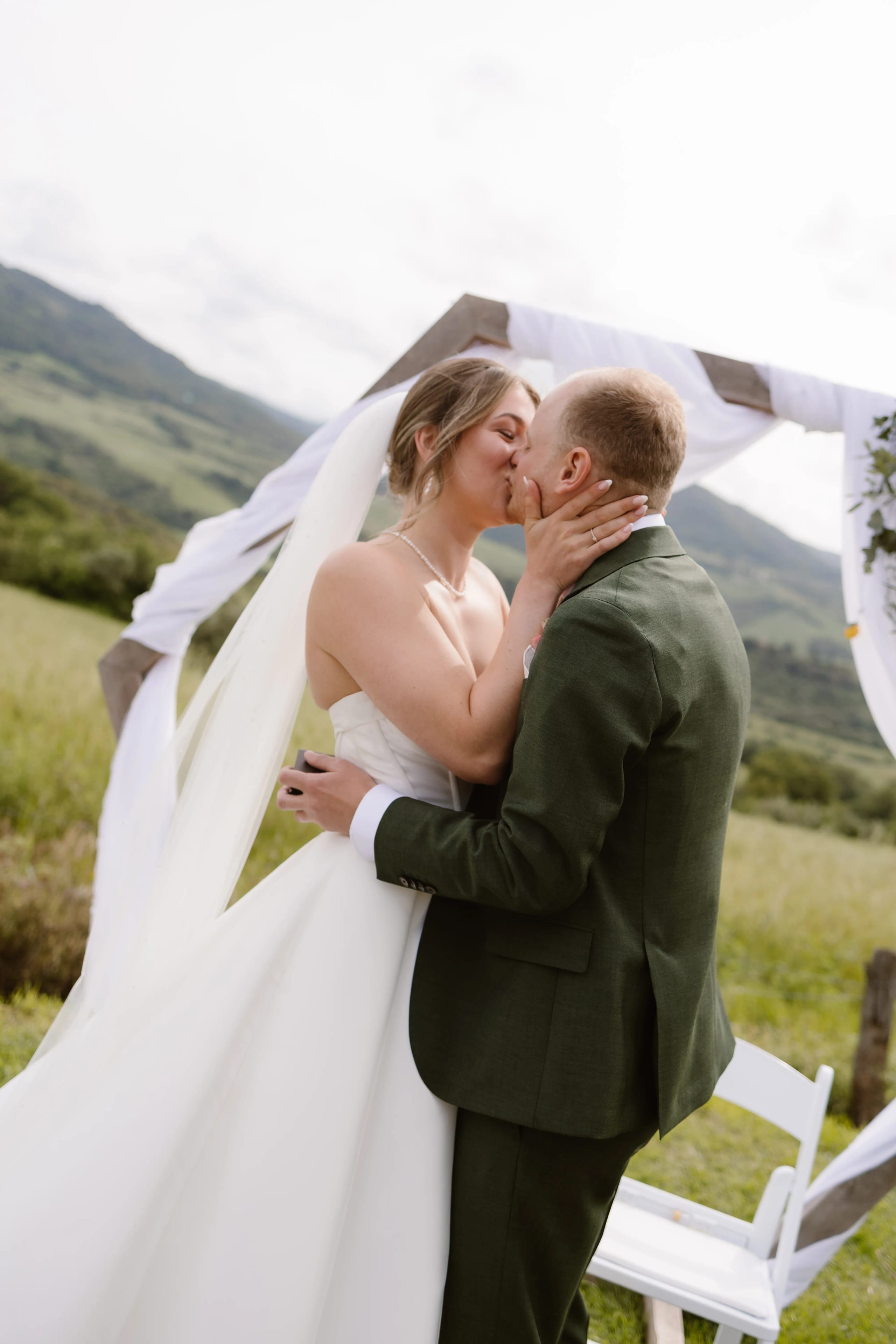 Een net getrouwd stel deelt een romantische kus onder een prachtig versierde trouwboog, omringd door groene heuvels en een serene natuurlijke omgeving. De bruid draagt een elegante jurk en de bruidegom een stijlvolle groene suit, wat de liefdevolle sfeer van deze onvergetelijke huwelijksceremonie accentueert.