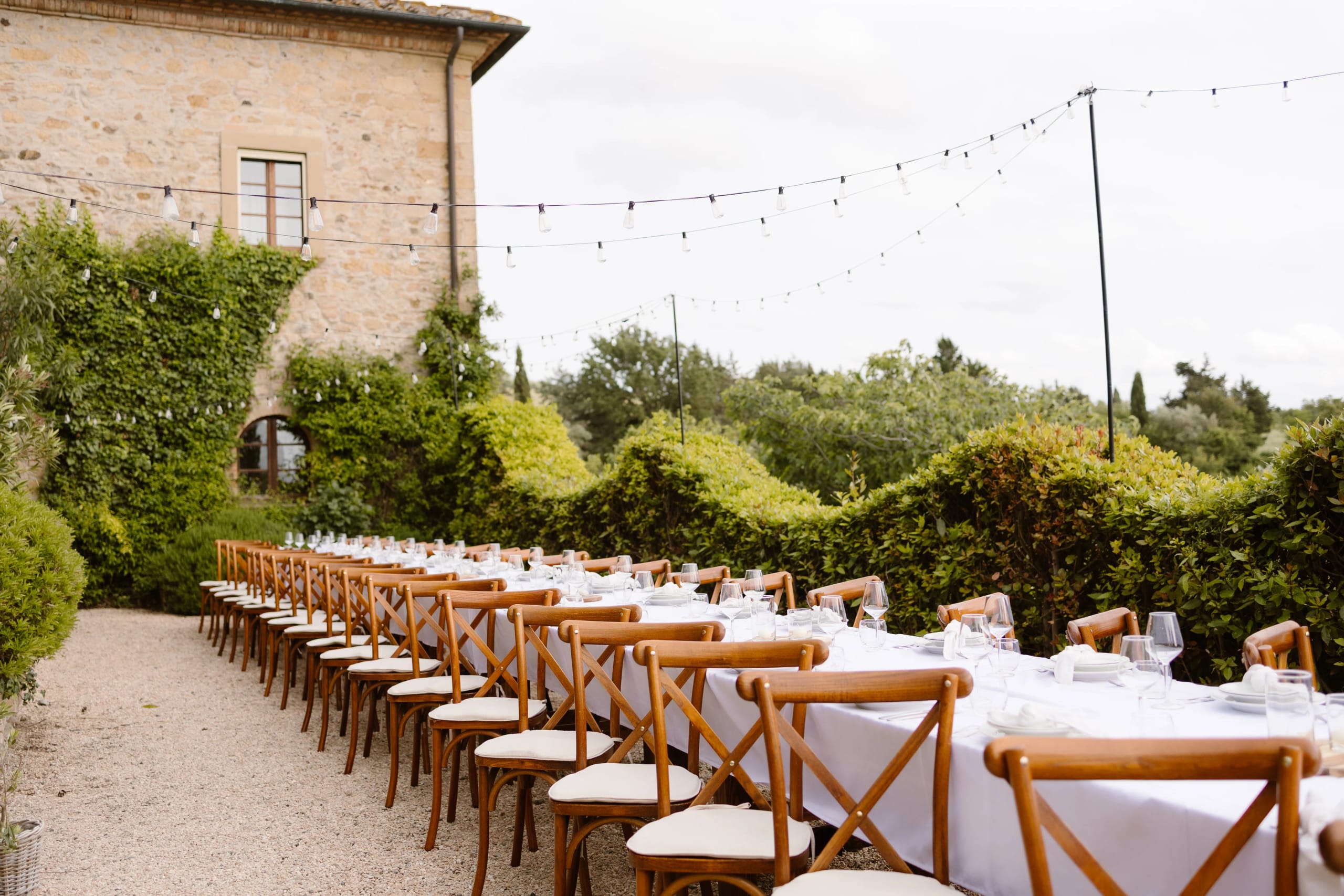 Een prachtig ingericht buitenrestaurant met lange, witte tafels en houten stoelen, omringd door weelderige groene begroeiing en sfeervolle hanglampen, ideaal voor een feestelijke gelegenheid of evenement.