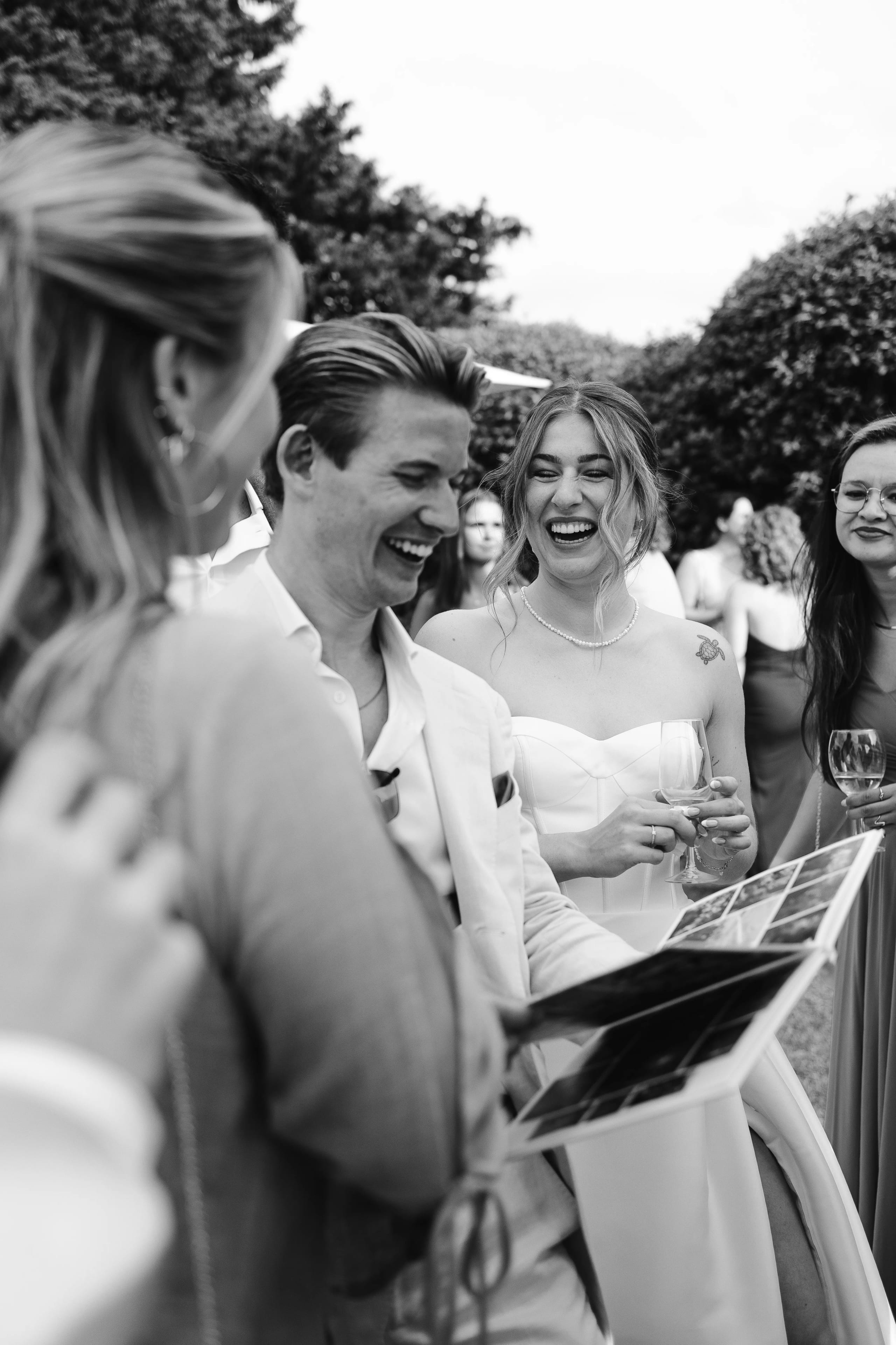 Een groep vrienden viert samen een gelukkige gelegenheid, met een lachende bruid in een witte trouwjurk die een gastenboek bekijkt, omringd door vrolijke gasten die genieten van een ontspannen buitenomgeving.
