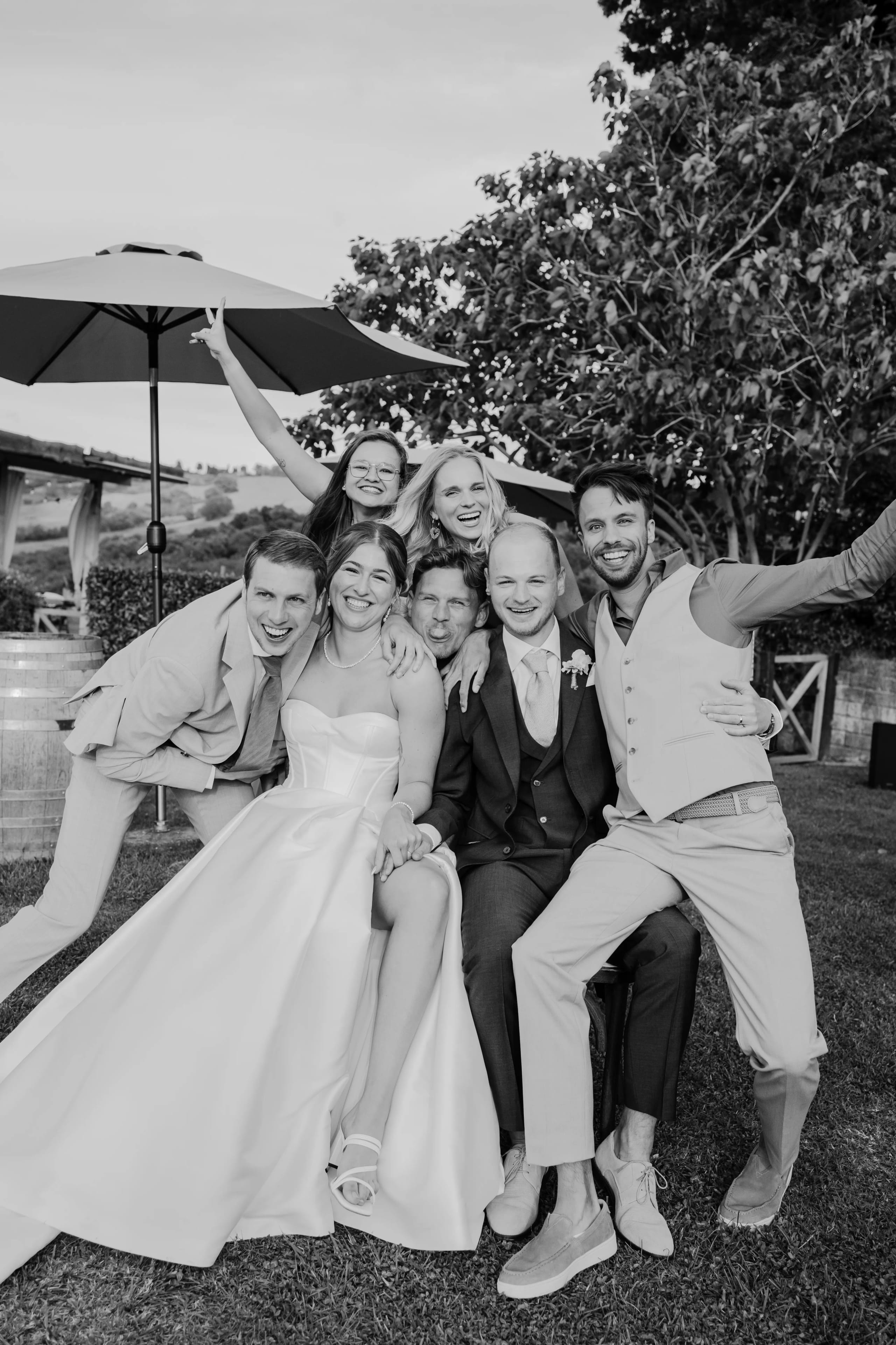 Zes vrienden poseren vrolijk samen, gekleed in formele kleding en een bruidsjurk, terwijl ze genieten van een zonnige buitenlocatie op een bruiloft met een parasol op de achtergrond.