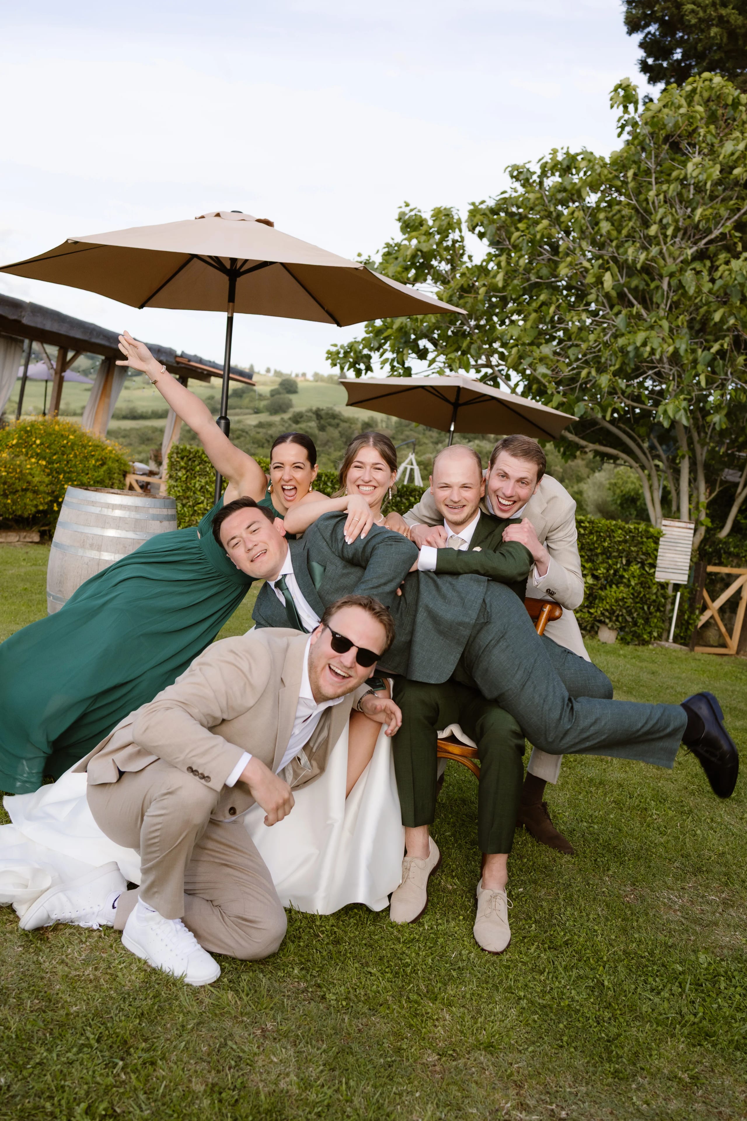 Een groep vrienden poseert vrolijk op een bruiloft in een groene tuin, met parasols op de achtergrond. Ze stralen gelach en plezier uit terwijl ze verschillende poses aannemen, gekleed in elegante outfits en feestelijke kleding.