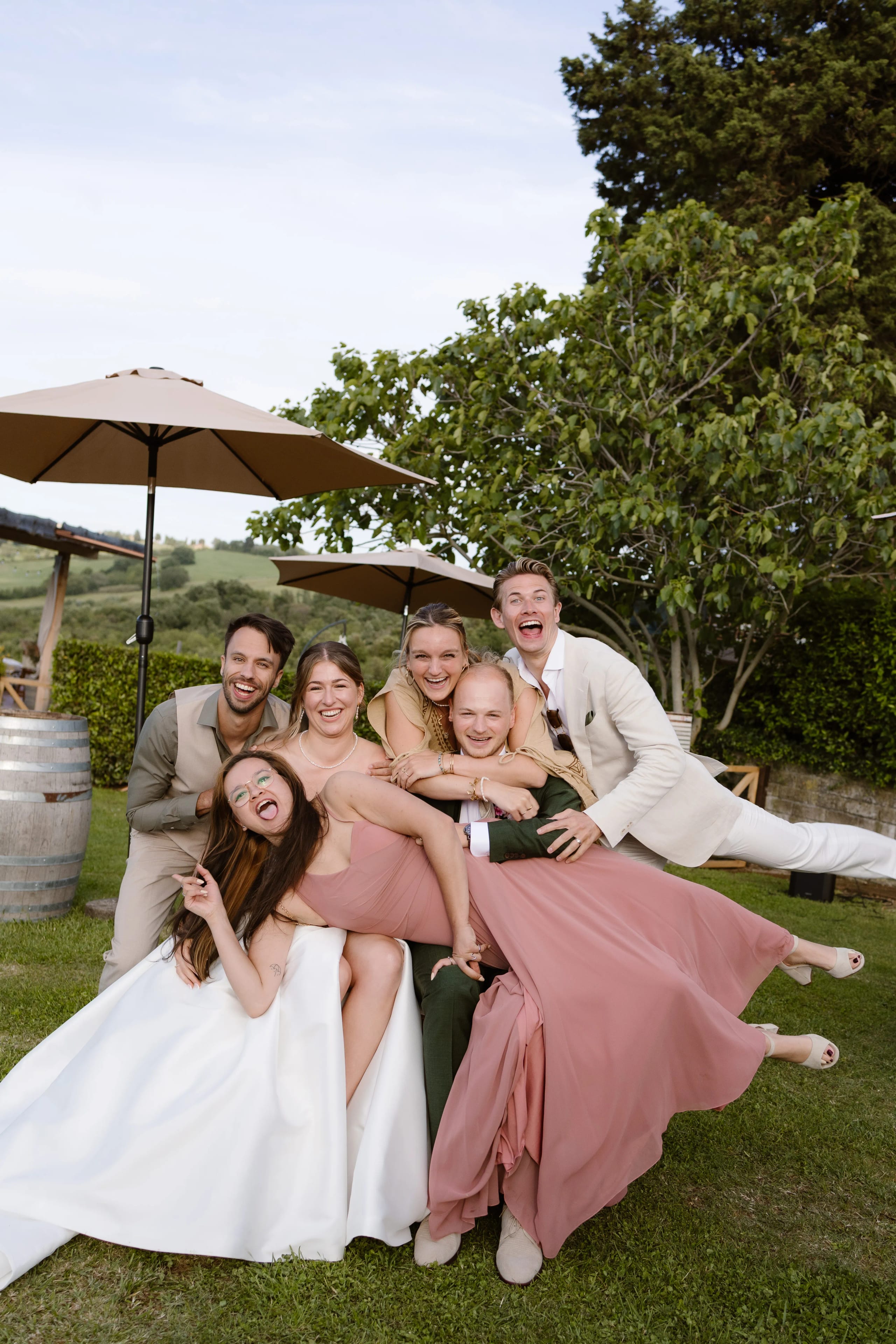 Een groep van zes vrolijke bruiloftsgasten poseert op een groen gazon, omringd door parasols en een landelijke achtergrond. De dames zijn gekleed in elegante avondjurken in verschillende tinten, terwijl de heren hun outfits afstemmen met stijlvolle accessoires, en iedereen straalt van blijdschap.