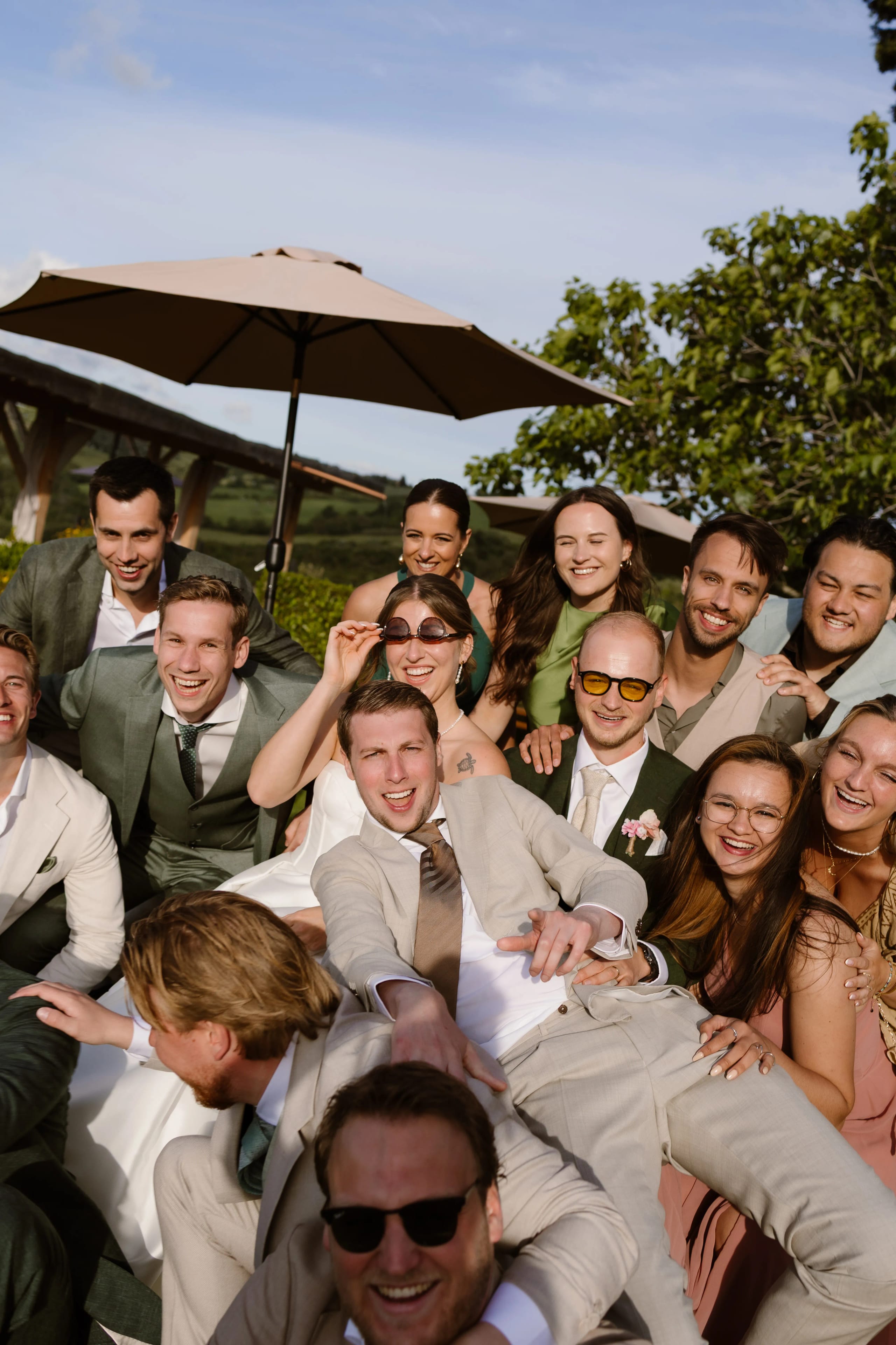 Een vrolijke groep van ongeveer vijftien mensen poseert samen op een zonnige locatie, omringd door groen, terwijl ze lachen, met enkele mensen in stijlvolle trouwoutfits en anderen in casual kleding. Achter hen staat een grote parasol die schaduw biedt, en het geheel straalt een gevoel van blijdschap en saamhorigheid uit.