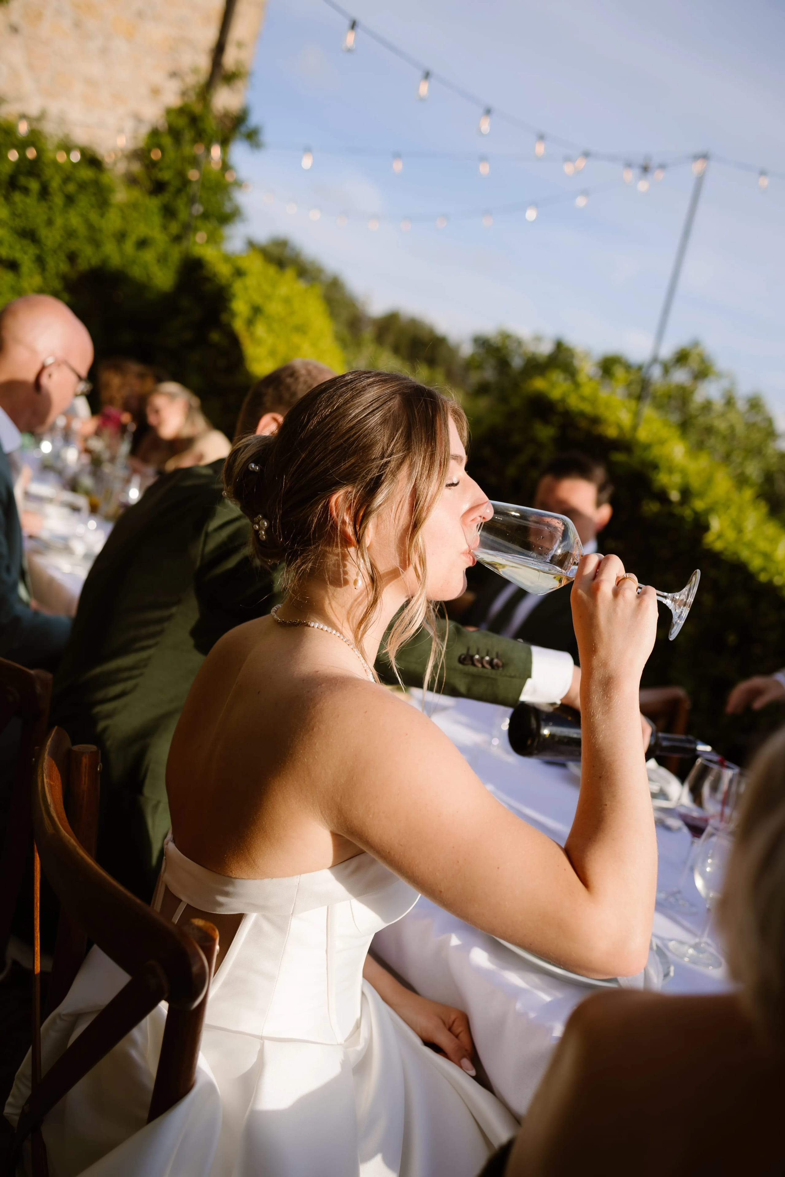 Een bruid in een witte trouwjurk geniet van een glas champagne tijdens een buitenbruiloft, omringd door gasten die achter een elegant gedekte tafel zitten, met sfeervolle verlichting en een groene achtergrond.