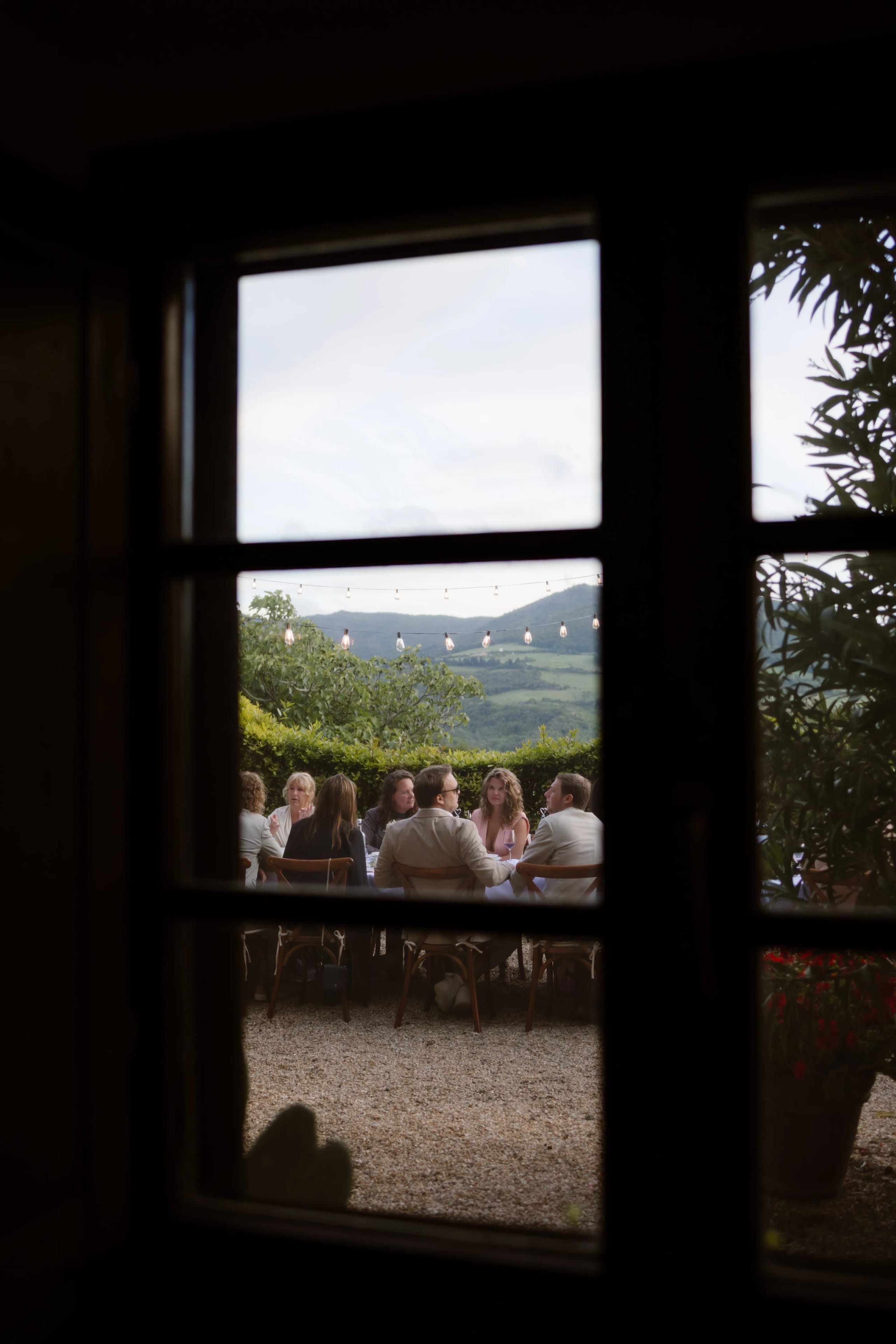 Gezellige groep mensen die samen aan een tafel dineren op een terras met uitzicht op groen heuvelachtig landschap, verlicht door sfeervolle lichtjes in de avondlucht.