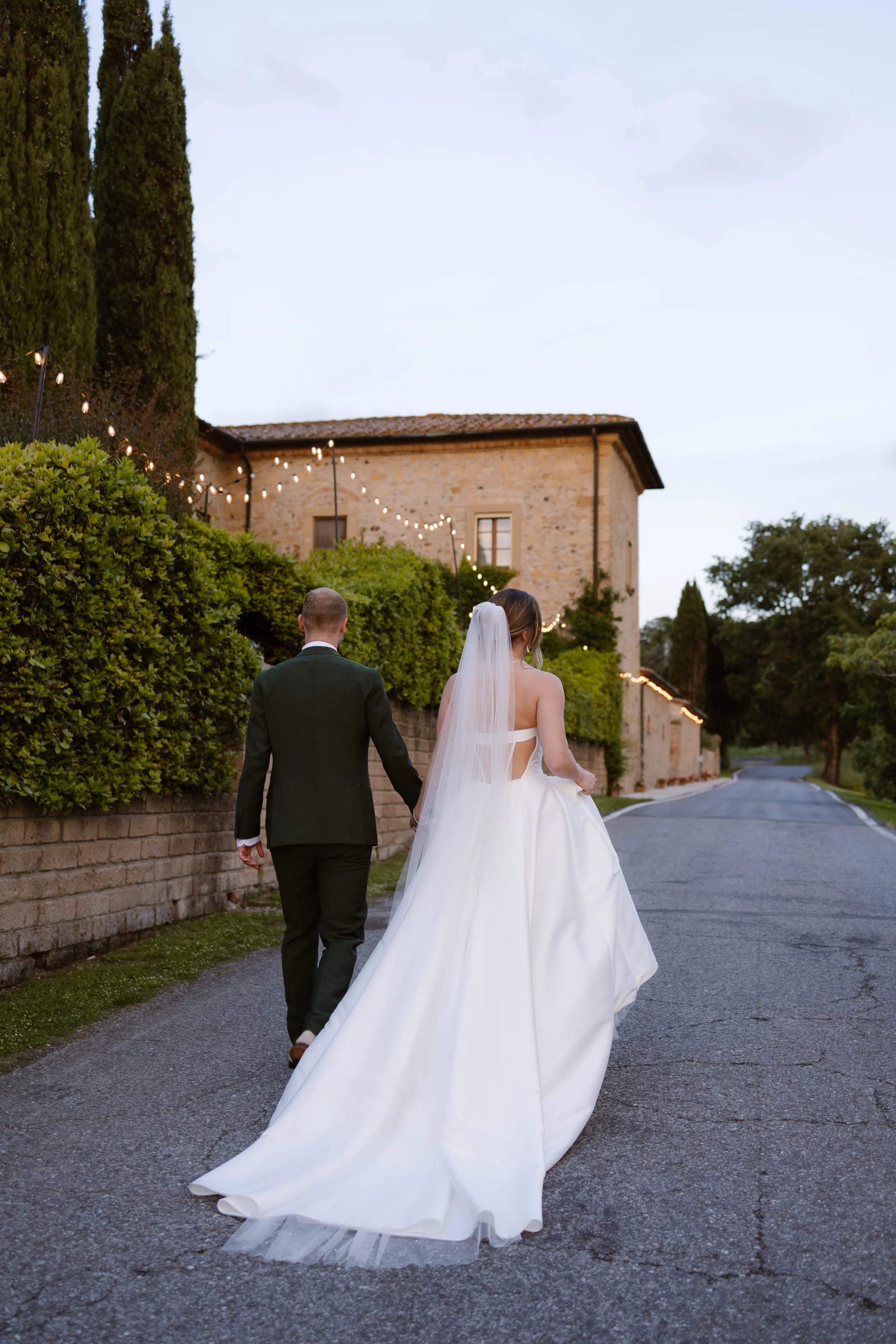Een net getrouwd stel wandelt hand in hand over een rustige, geplaveide weg omringd door groene hagen en sfeerverlichting, met een prachtig landhuis op de achtergrond, perfect voor een romantische fotoshoot.