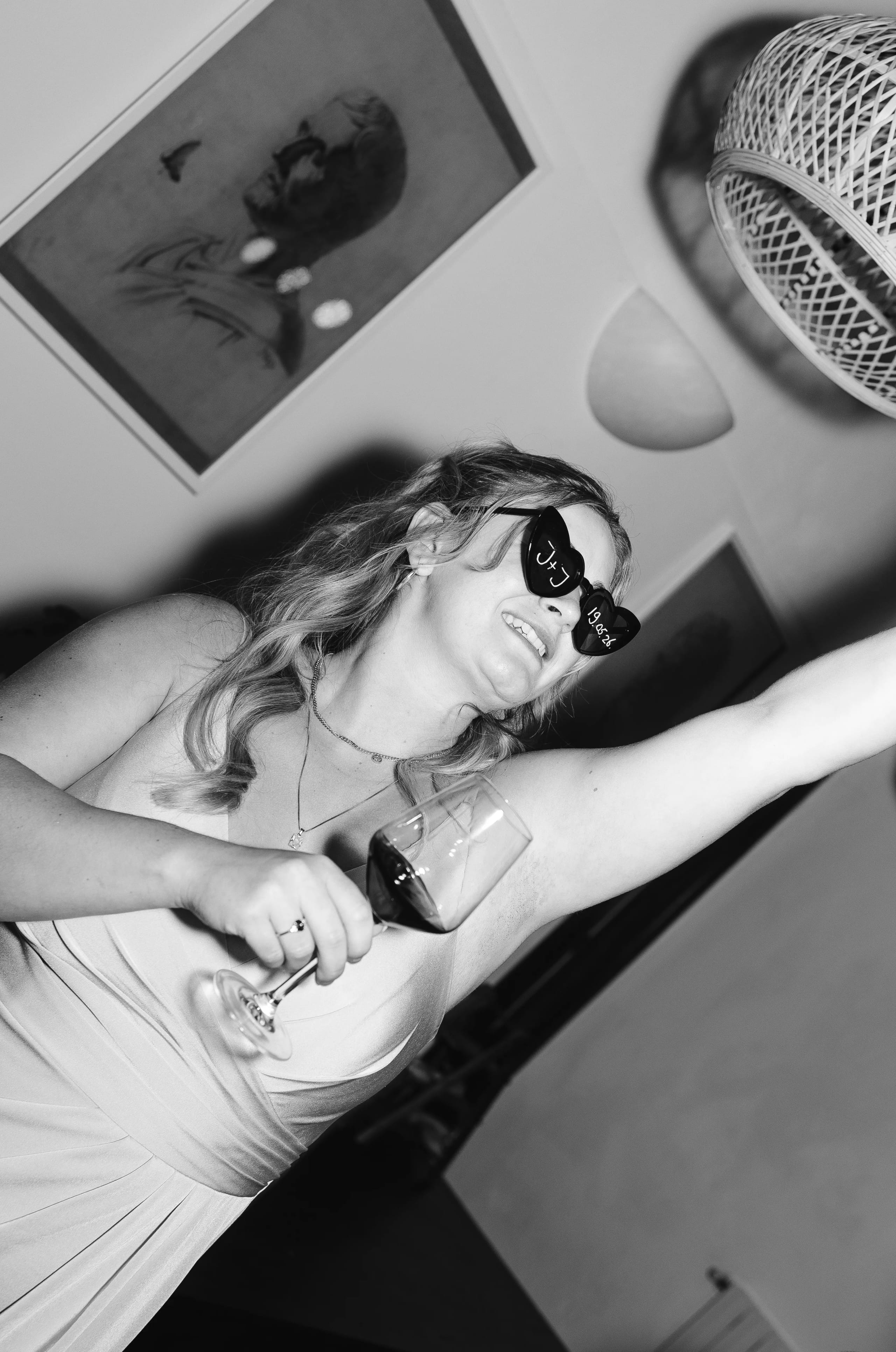 Een vrouw in een elegante jurk, met een glas wijn in de hand en een brede glimlach, danst vrolijk terwijl ze met haar andere hand naar boven wijst, omringd door een sfeervolle interieursetting.
