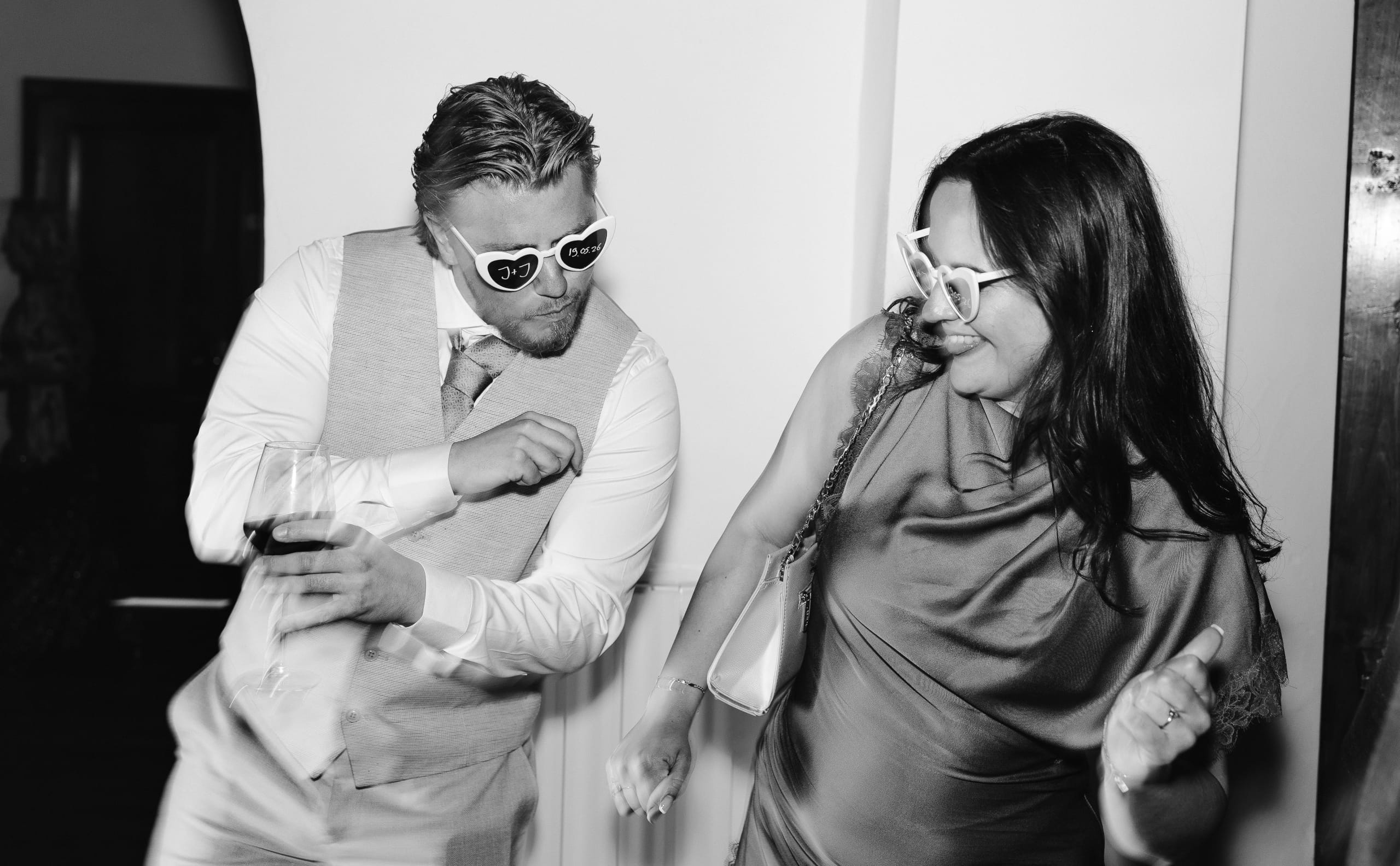 Een man en een vrouw dansen enthousiast op een feest, beiden met kleurrijke feestbrillen op. De man draagt een lichtgrijze vest en houdt een glas in zijn hand, terwijl de vrouw in een elegante outfit met een handtas aan haar schouder glimlacht en meedenkt in de dans.