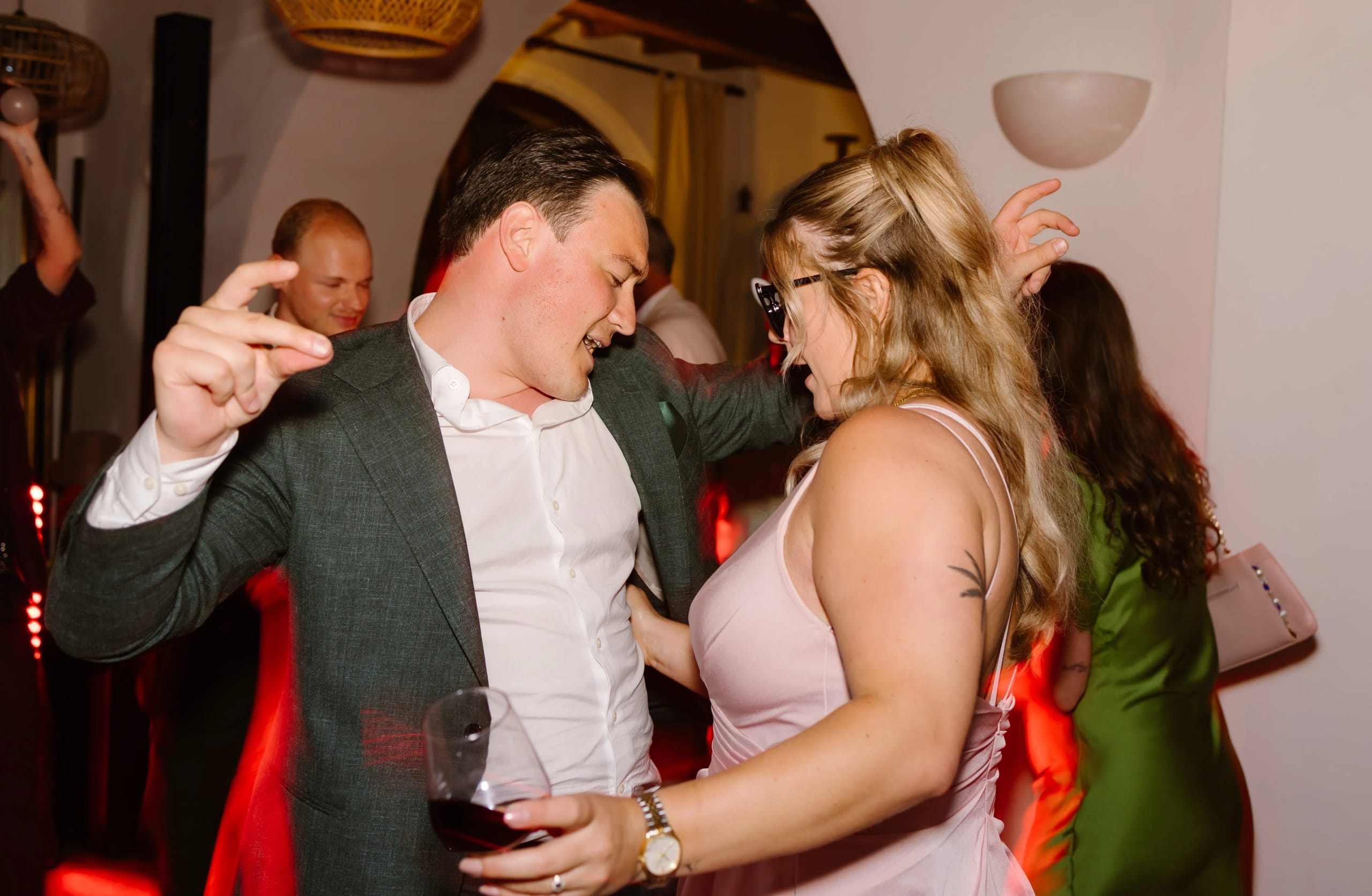 Een man in een stijlvolle blazer danst enthousiast met een vrouw in een roze top, terwijl ze beide genieten van een levendige feestelijke sfeer met sfeerverlichting op de achtergrond. De vrouw houdt een glas rode wijn vast.
