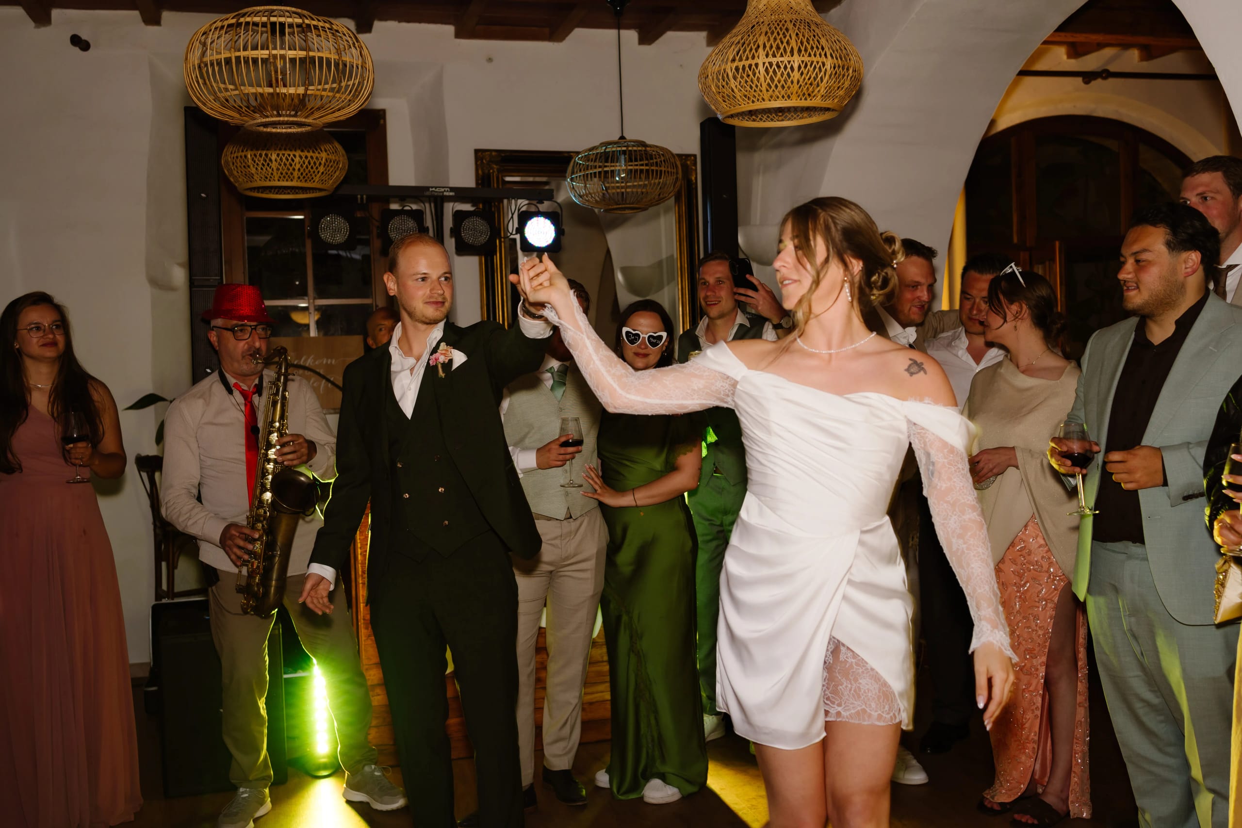 Een bruid in een moderne, schijnt met een zaklamp terwijl ze omringd wordt door feestvierende gasten in een sfeervolle, goed verlichte ruimte, met decoratieve rieten lampen en muziekanten op de achtergrond.