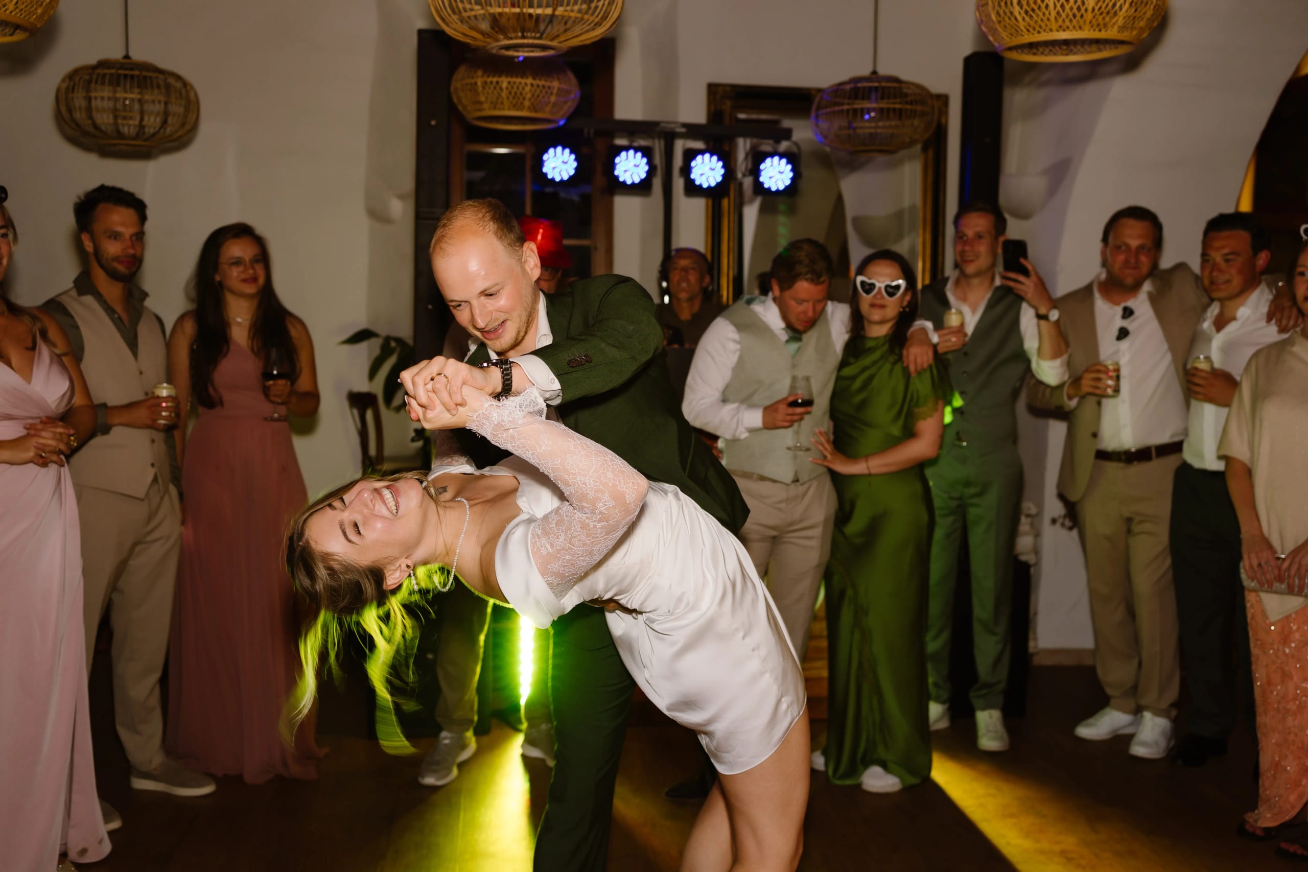 Een man in een groen kostuum danst met een vrouw in een witte jurk, terwijl ze een elegante draaibeweging maakt. Achter hen zijn gasten in feestelijke kleding te zien, die enthousiast kijken en genieten van de sfeer op het dansfeest.
