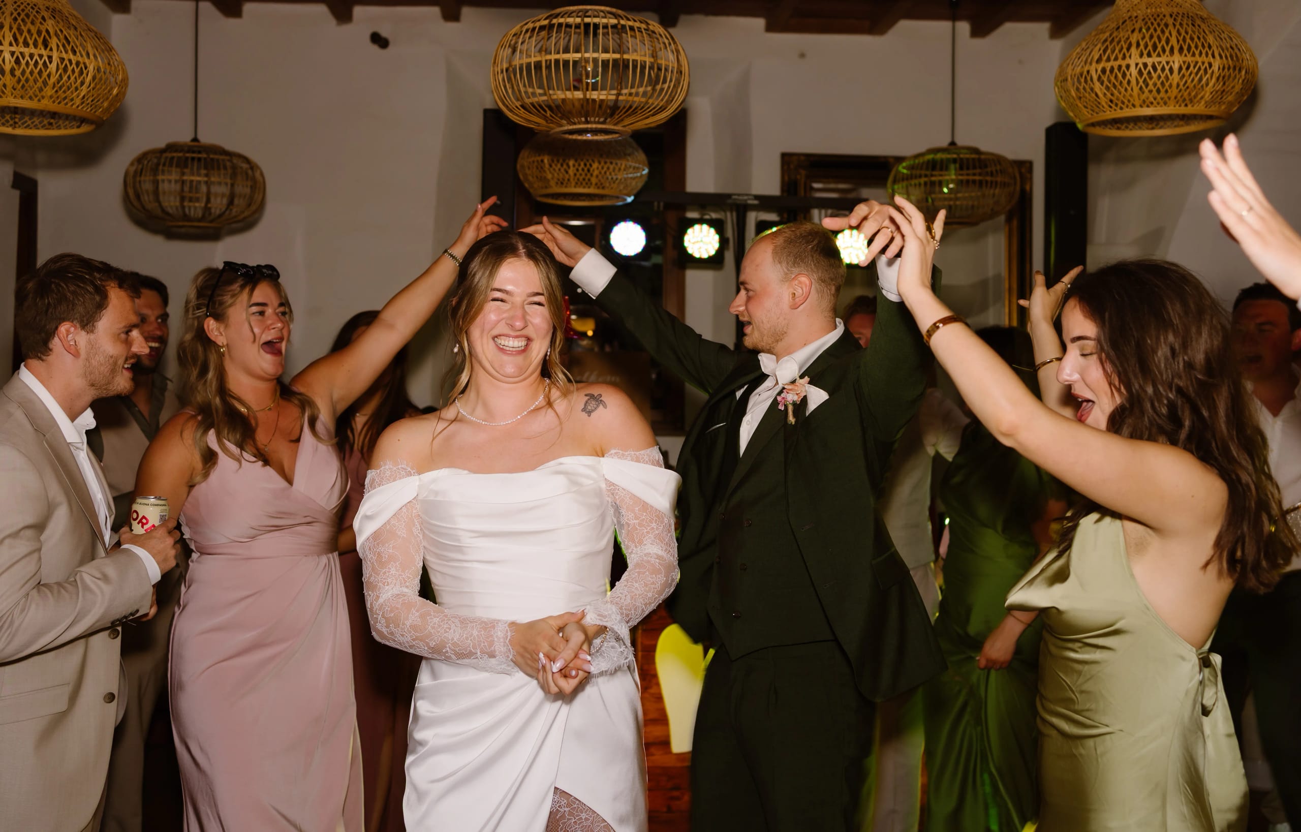 Een vrolijke bruid in een stralende witte jurk danst met vrienden, omringd door blije gasten die hun handen in de lucht steken tijdens een feestelijke gelegenheid met sfeervolle verlichting en een gezellige ambiance.