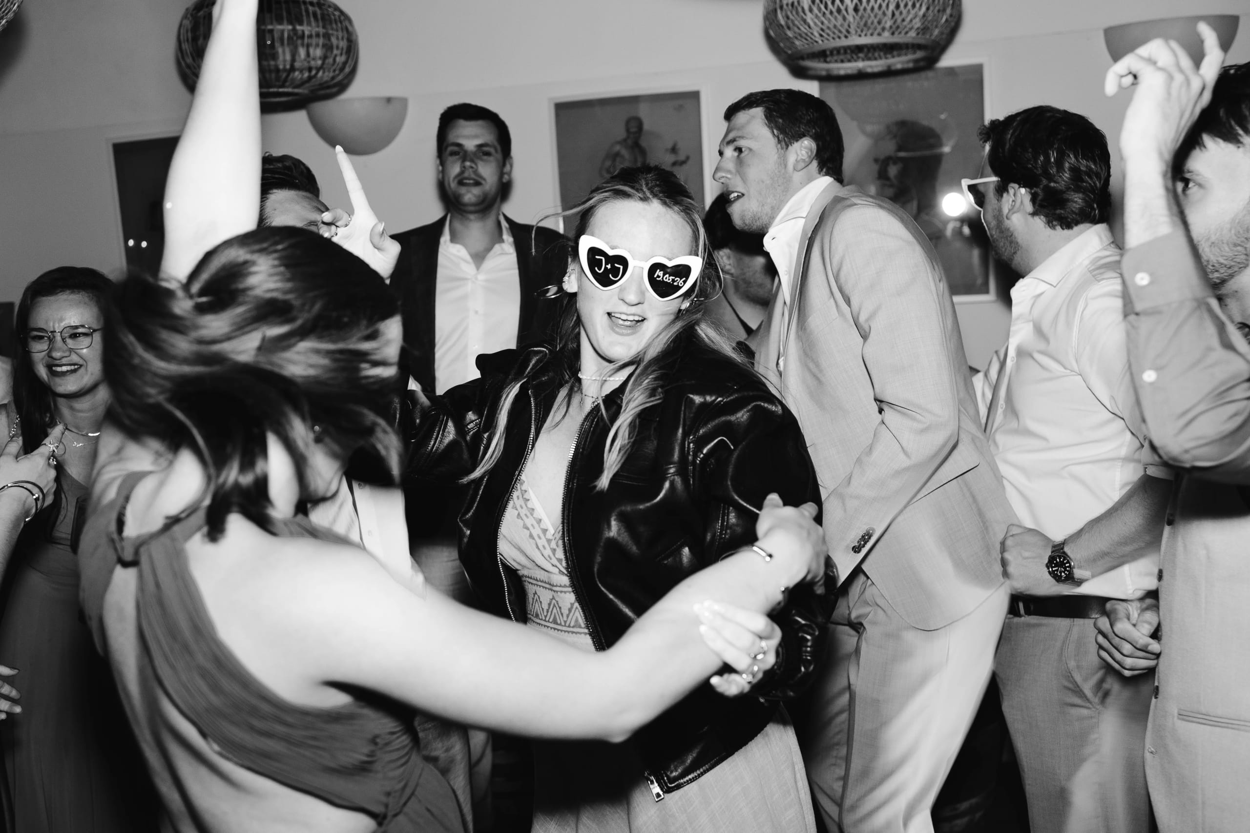 Een energieke dansscene met feestende gasten in een fraaie setting; een vrouw in een zwarte leren jack en hartvormige zonnebril danst vrolijk met een andere vrouw, terwijl de opzwepende sfeer wordt versterkt door andere feestgangers op de achtergrond.