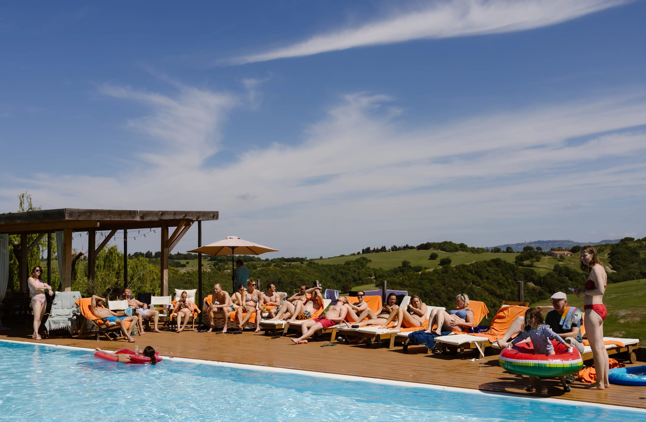 Een levendige zomerse scène bij een zwembad met meerdere mensen die ontspannen op ligstoelen liggen, genieten van de zon onder een heldere blauwe lucht, met een groene heuvelachtige achtergrond en een paar opblaasbare zwembadspeelgoed.