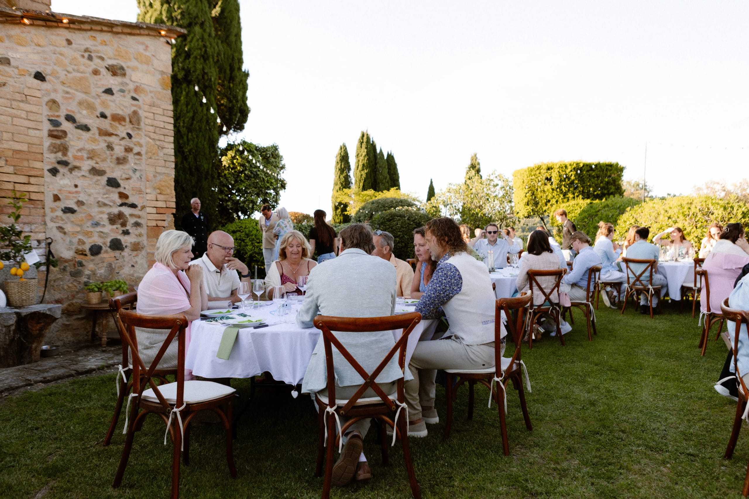 Een groep mensen geniet van een elegant diner in een prachtige tuin, omringd door groene planten en een historische stenen muur, met sfeervolle verlichting in de avondlucht.