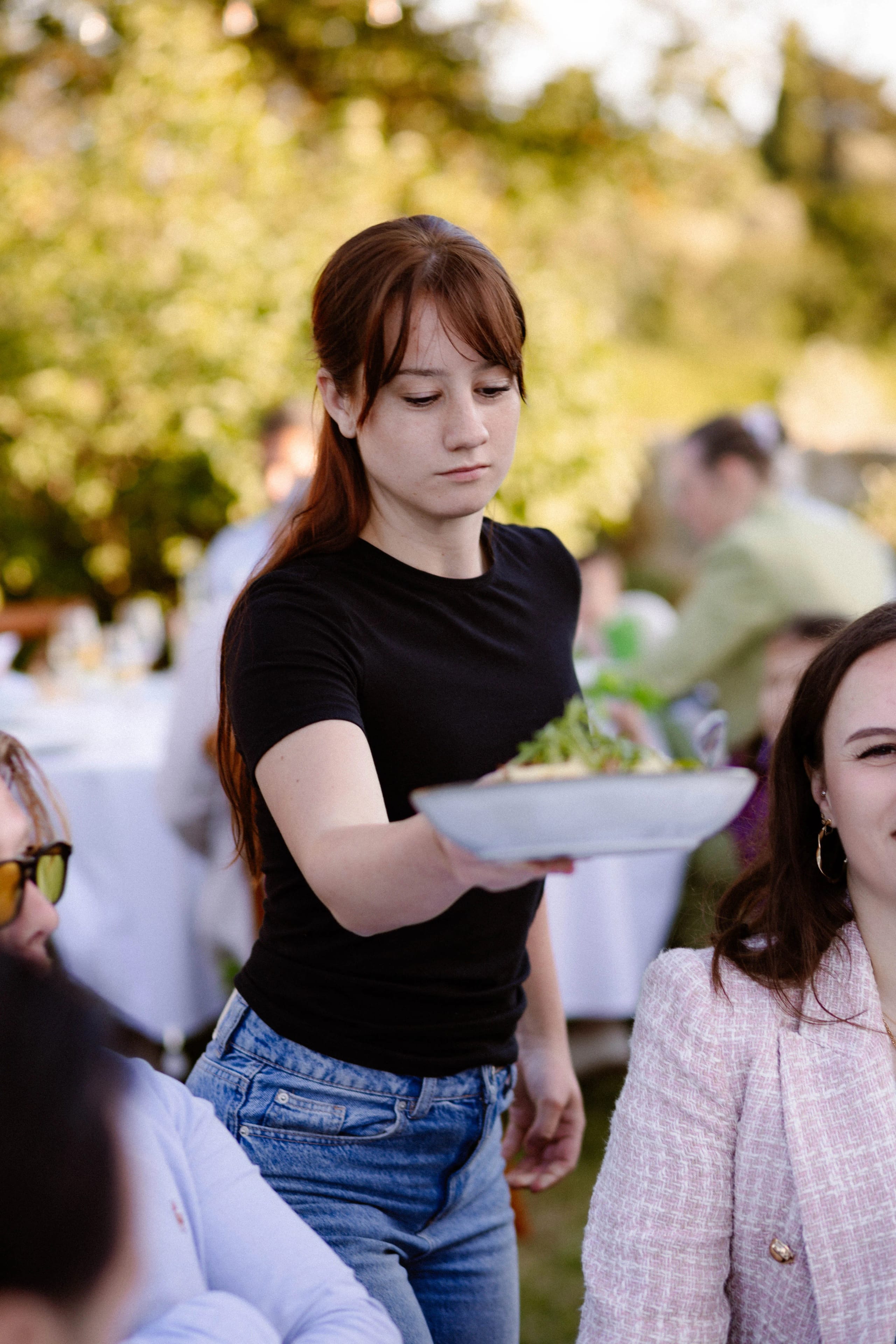 Een vrouw met lange kastanjebruine haren in een zwart T-shirt serveert een bord met salade tijdens een buitenbijeenkomst, terwijl gasten aan tafel in een gezellige sfeer deelnemen aan het evenement.